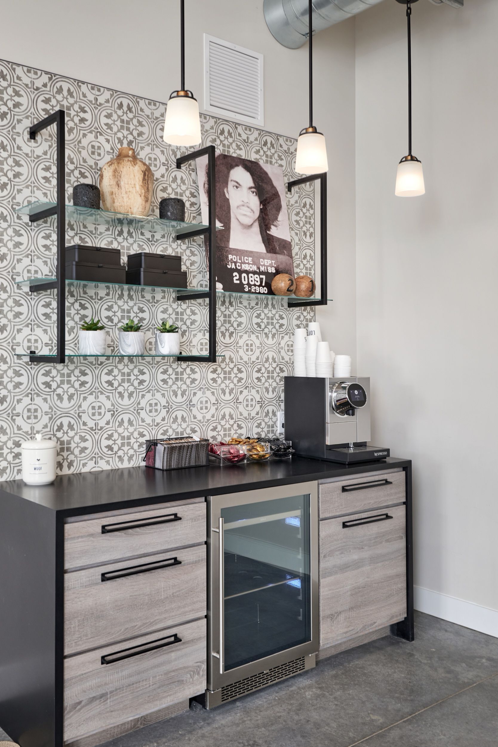 Modern kitchenette with black counter, mini fridge, wall shelves, and hanging pendant lights