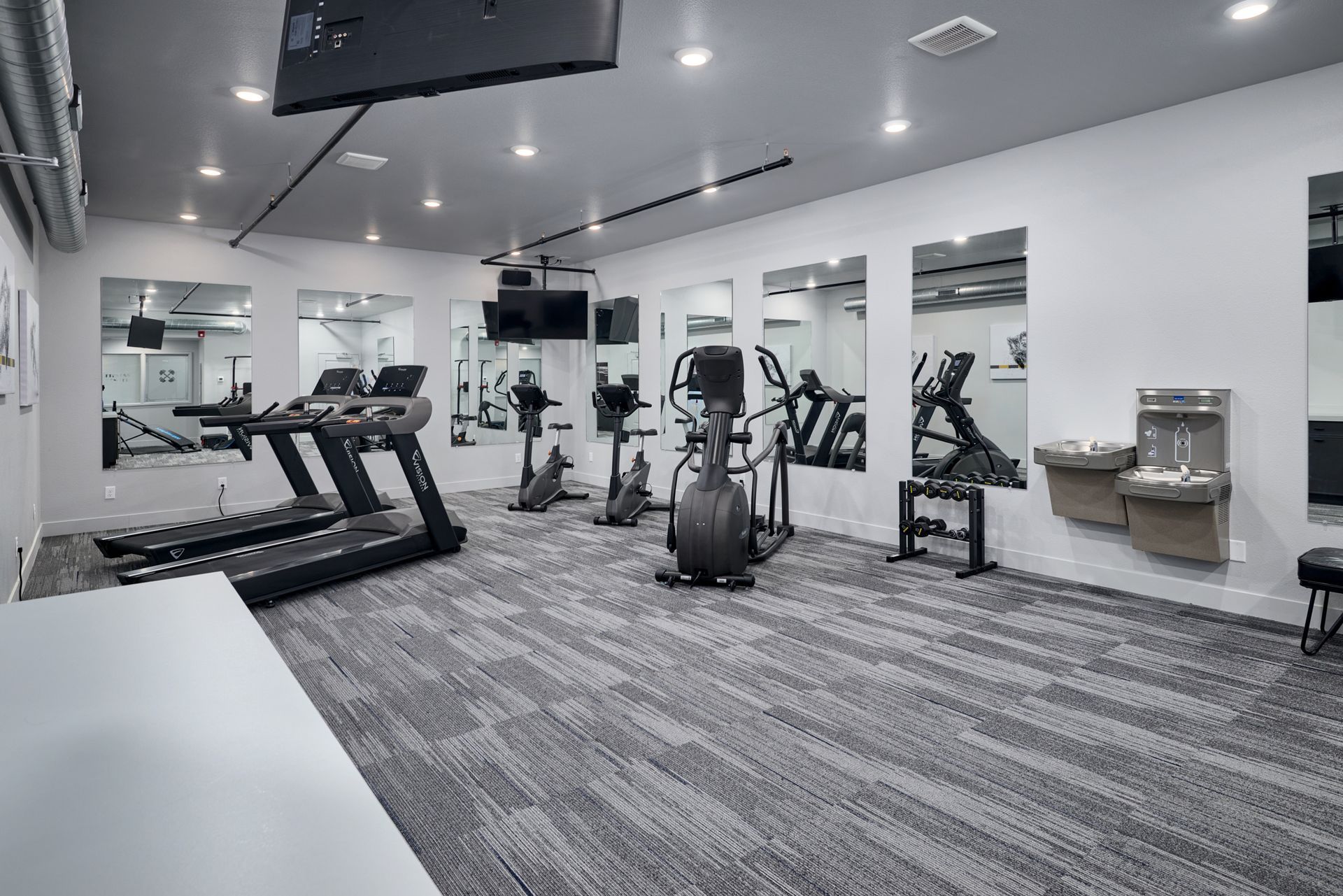 Empty modern gym with treadmills, ellipticals, strength machines, mirrors, and water fountain under bright lights