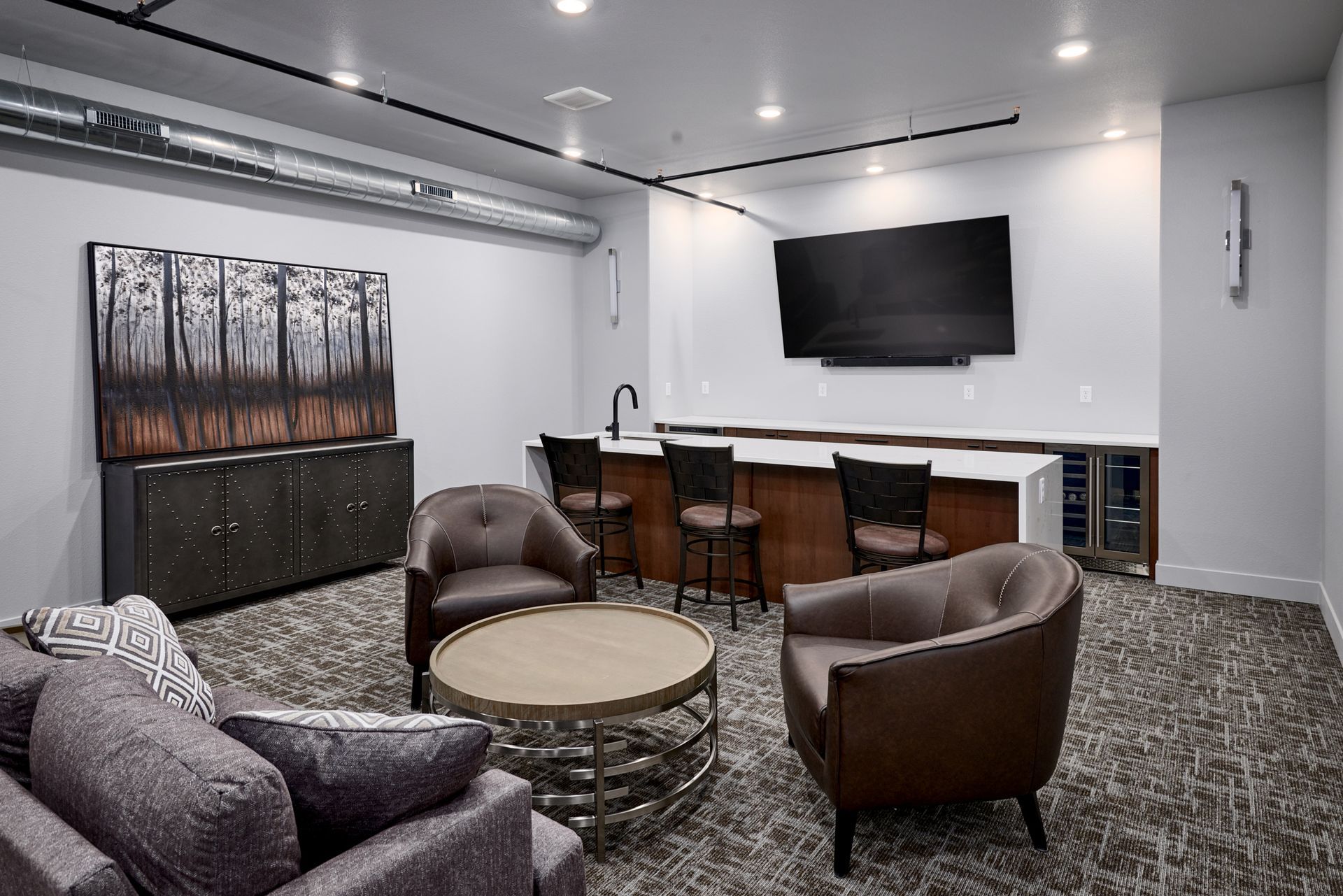Modern lounge with leather chairs, round table, bar stools, wall art, and a mounted TV under bright lights