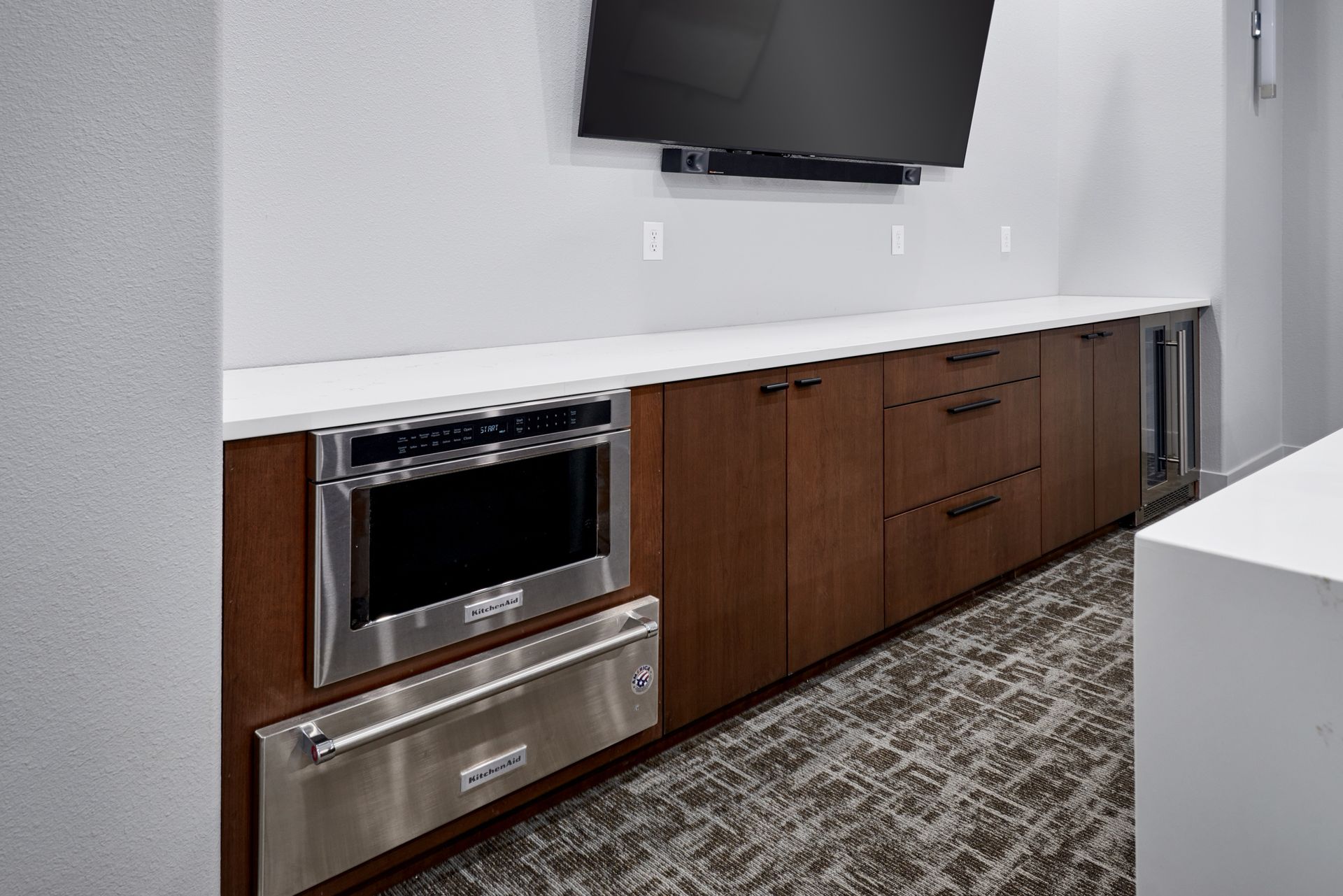 Modern kitchenette with wood cabinets, stainless oven, wall-mounted TV, and patterned carpet