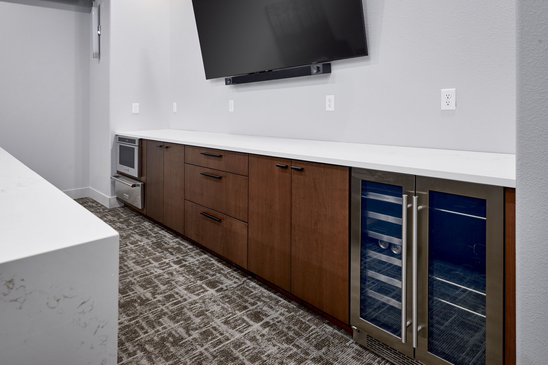 Modern break room with wall-mounted TV, long counter, wooden cabinets, and mini fridges along the wall.