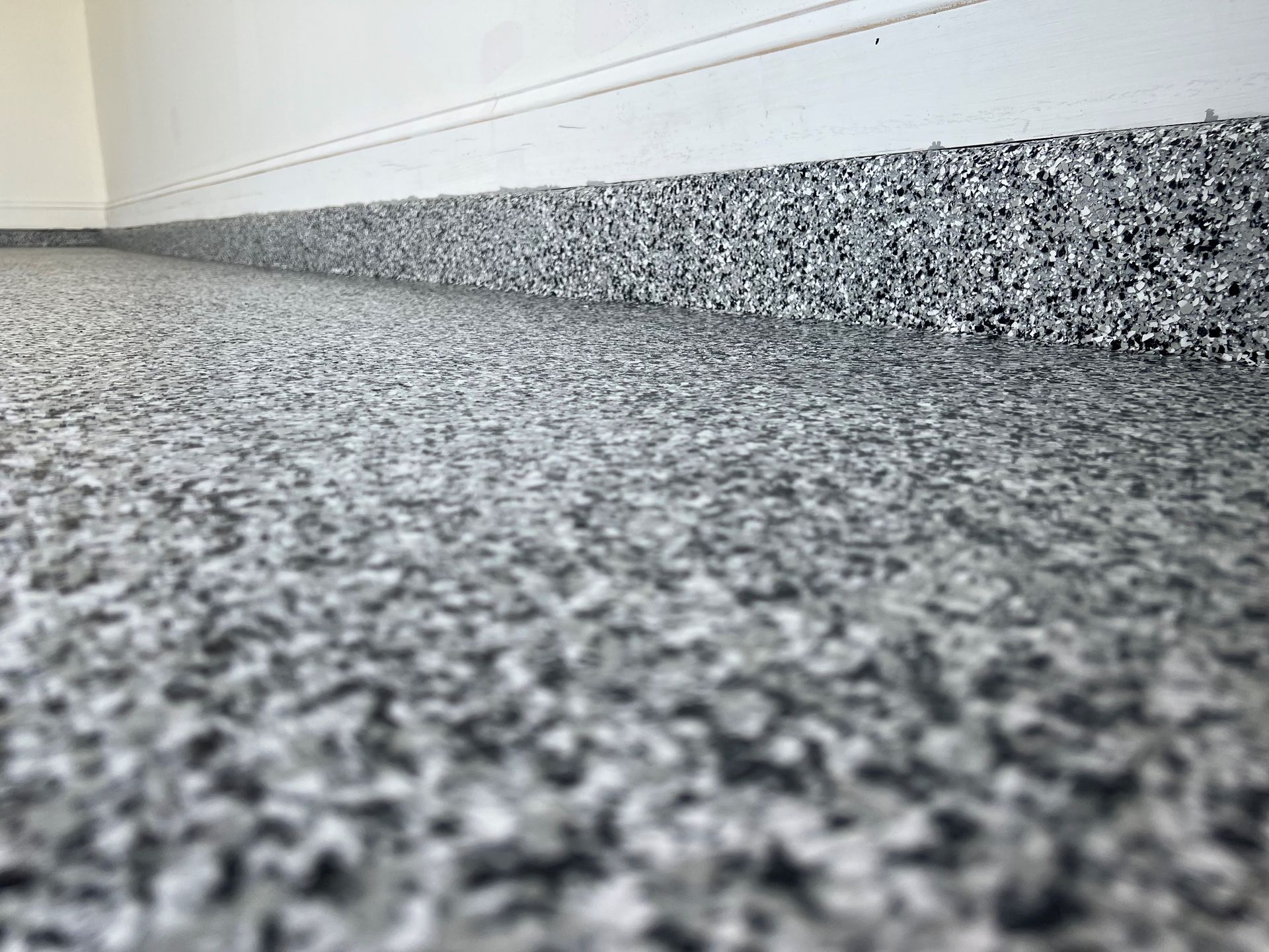 Gray speckled epoxy flooring in a garage, against white walls and a baseboard.