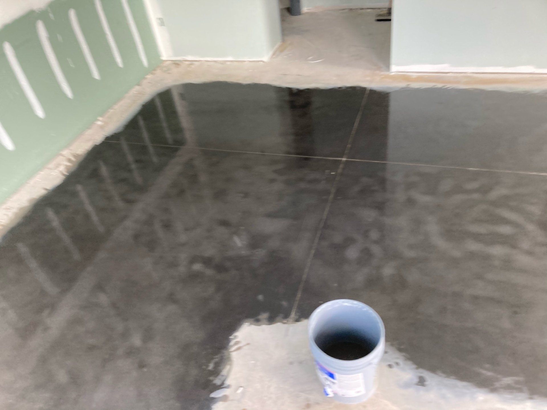 A wet, dark gray floor reflects light in a room under construction. A white bucket sits on the floor near a corner.