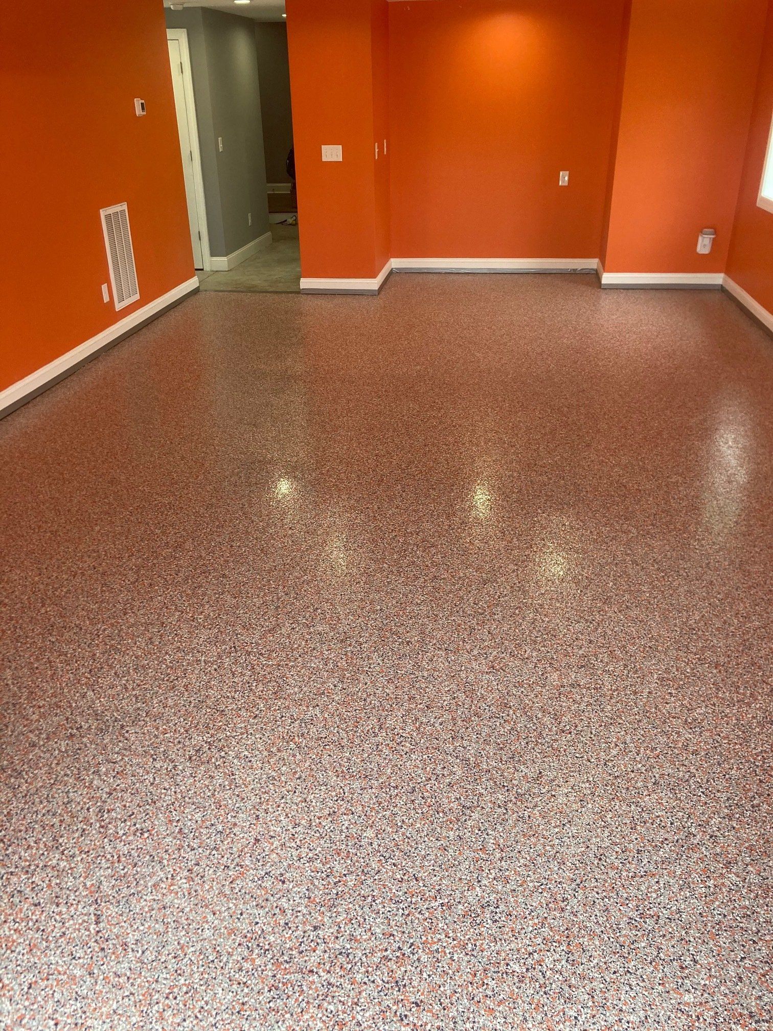 A room with a speckled red and white epoxy floor and orange walls. A doorway leads to a hallway with gray walls.
