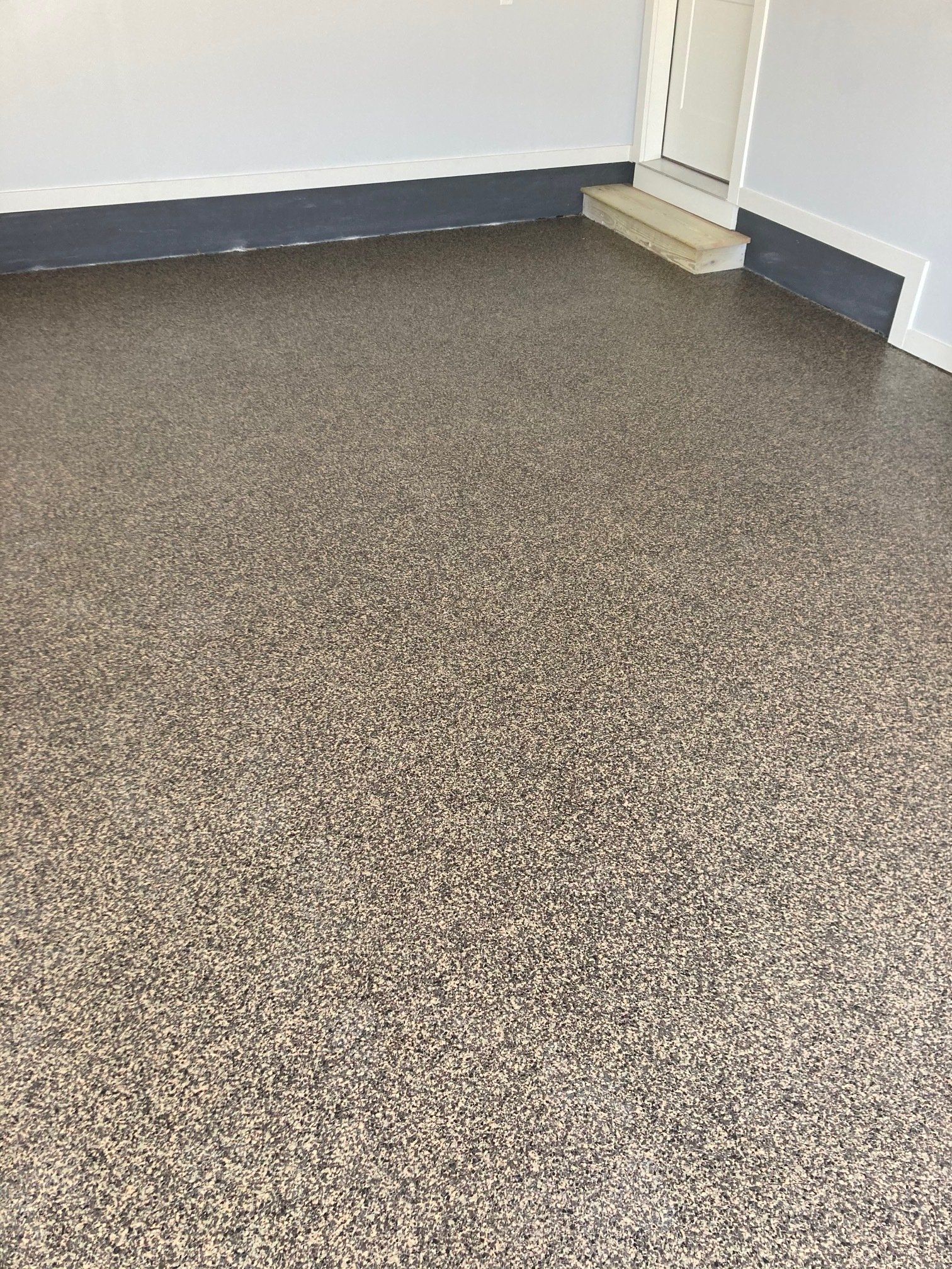 Garage floor with a speckled, neutral-toned epoxy coating. The walls are painted gray with a dark blue baseboard.