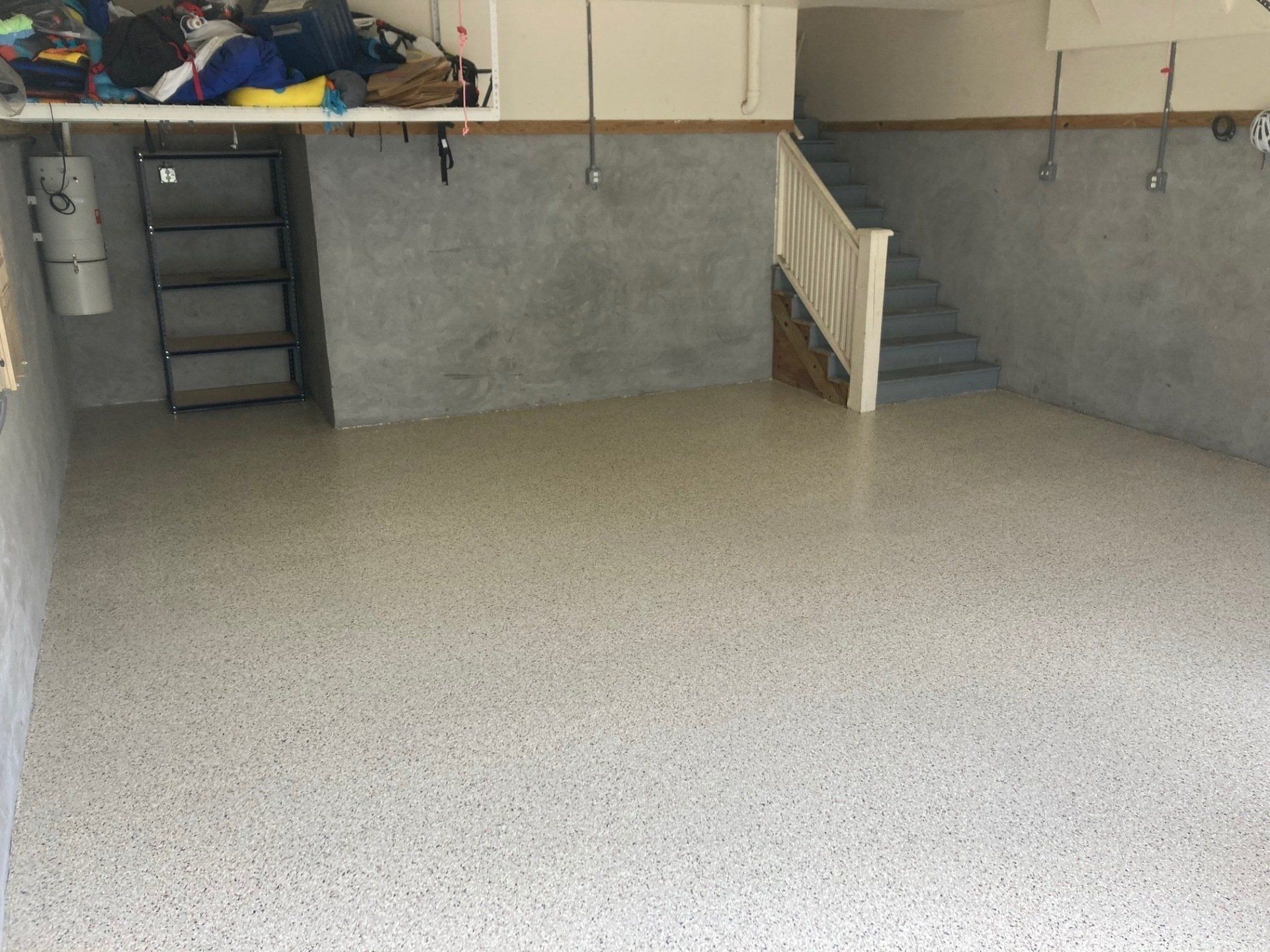 A clean, newly coated garage floor with a specked, light gray appearance. A staircase leads upward.