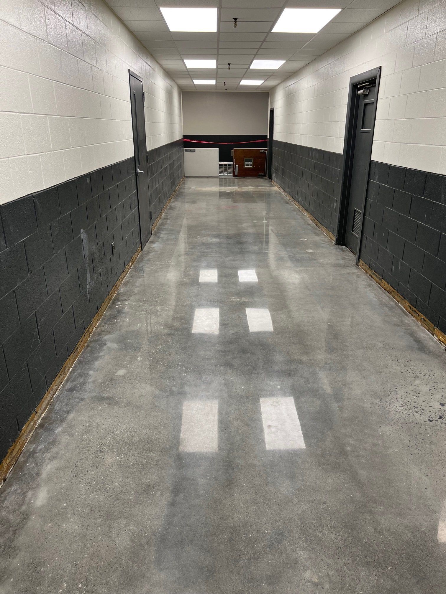 A narrow hallway with polished concrete floors and black and white block walls. Overhead, square lights reflect on the floor.