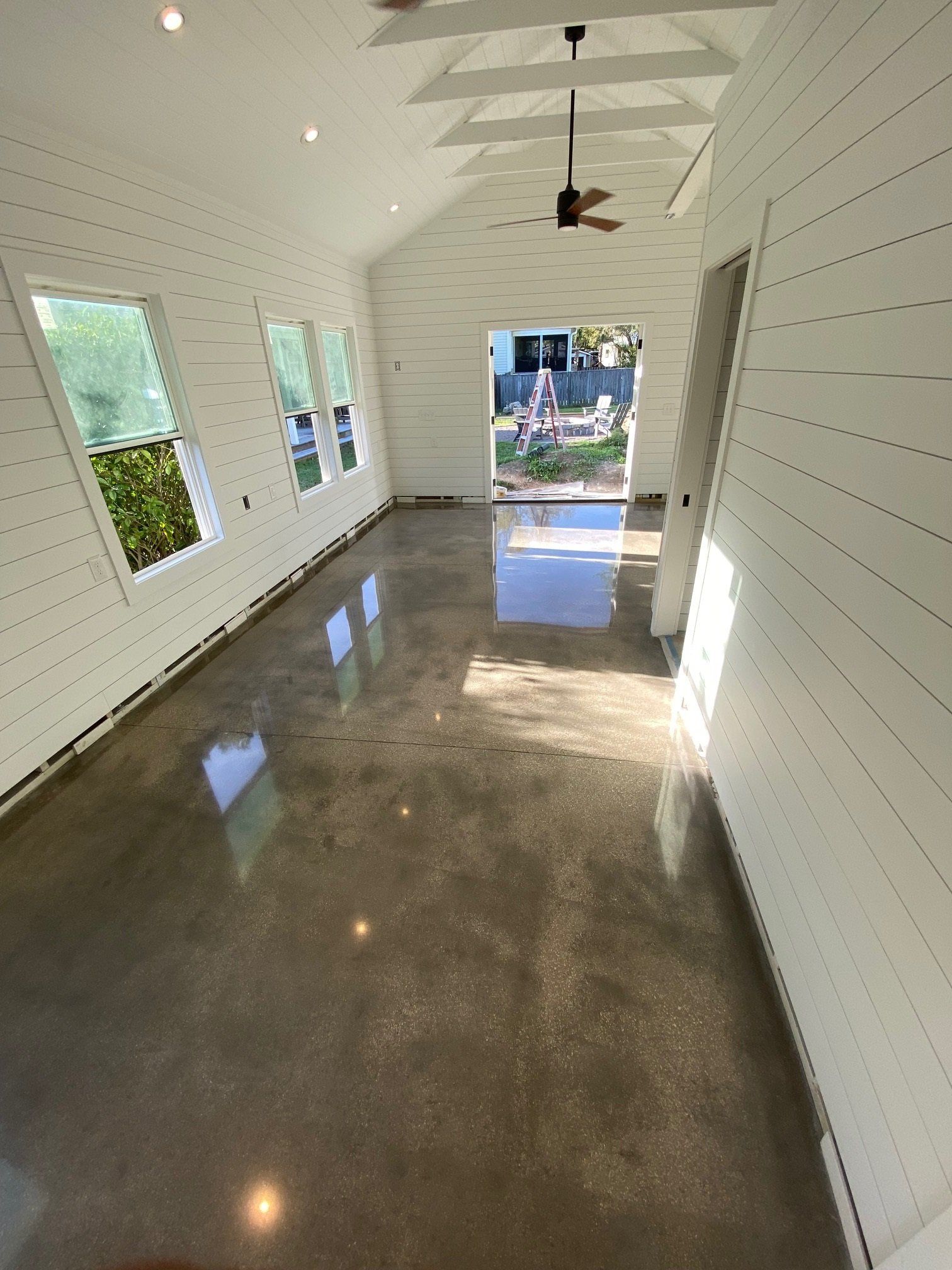 Bright interior with polished concrete floor, white shiplap walls, and three windows. An open doorway leads outside.