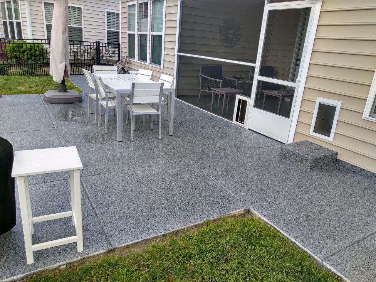 Outdoor patio with gray speckled concrete, white furniture, and a screened door. Green grass borders the patio.