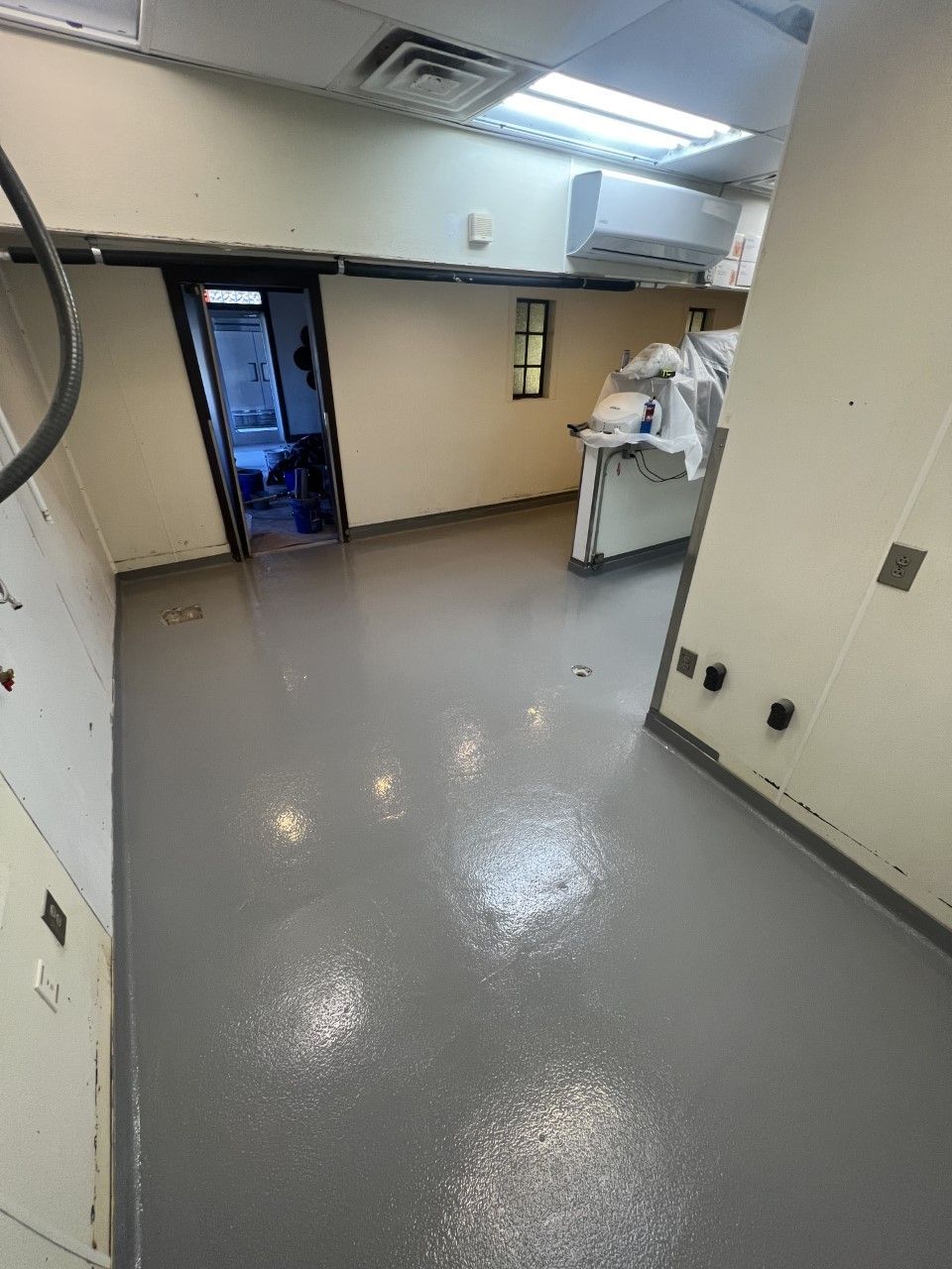 Empty commercial kitchen with gray epoxy floor, neutral walls, and a doorway leading to another space.