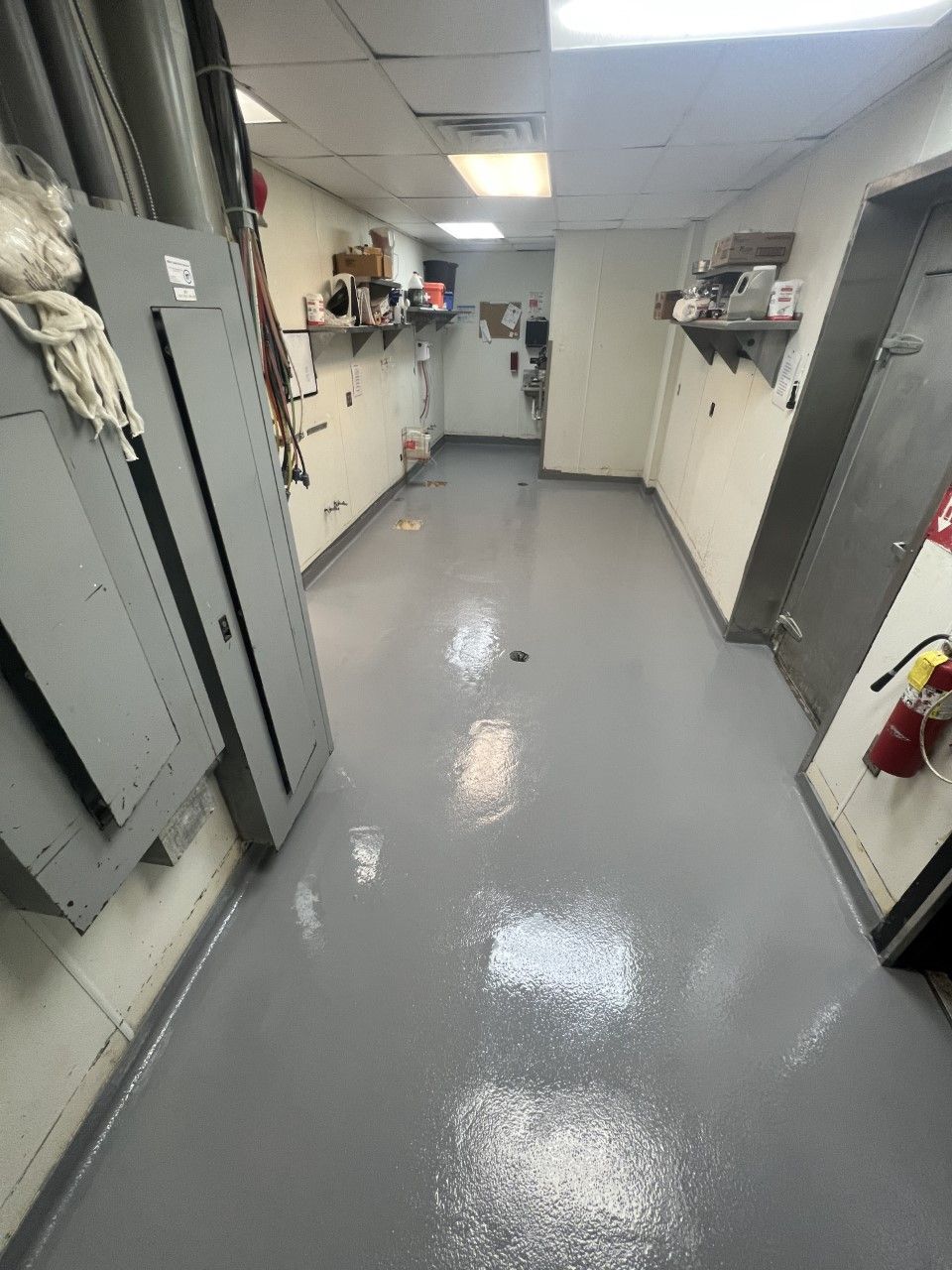 A narrow commercial hallway with gray painted floor and walls, featuring electrical panels, shelves, and closed doors.