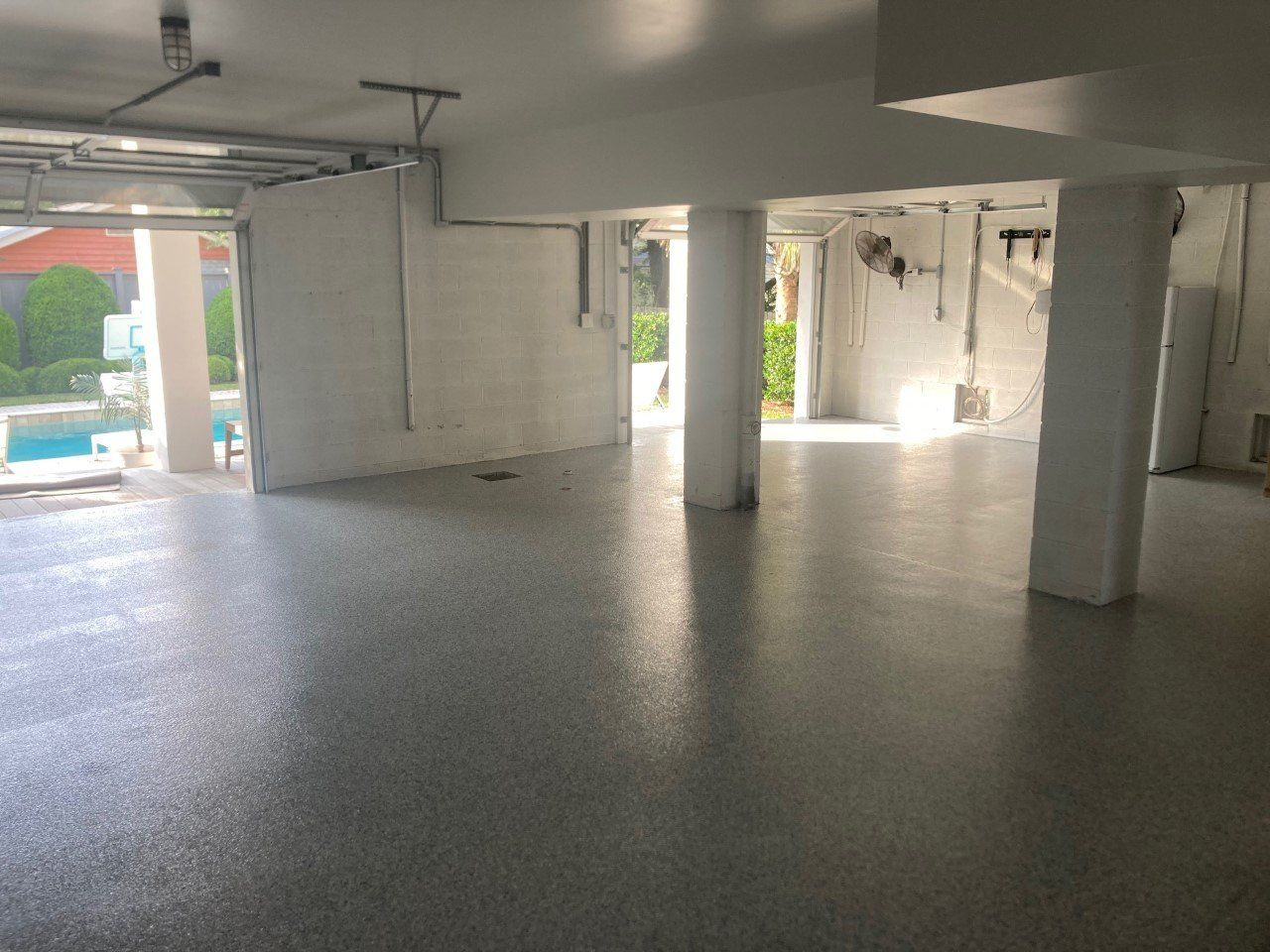 Empty garage with gray epoxy flooring, white columns, and a view of a pool through an open doorway.