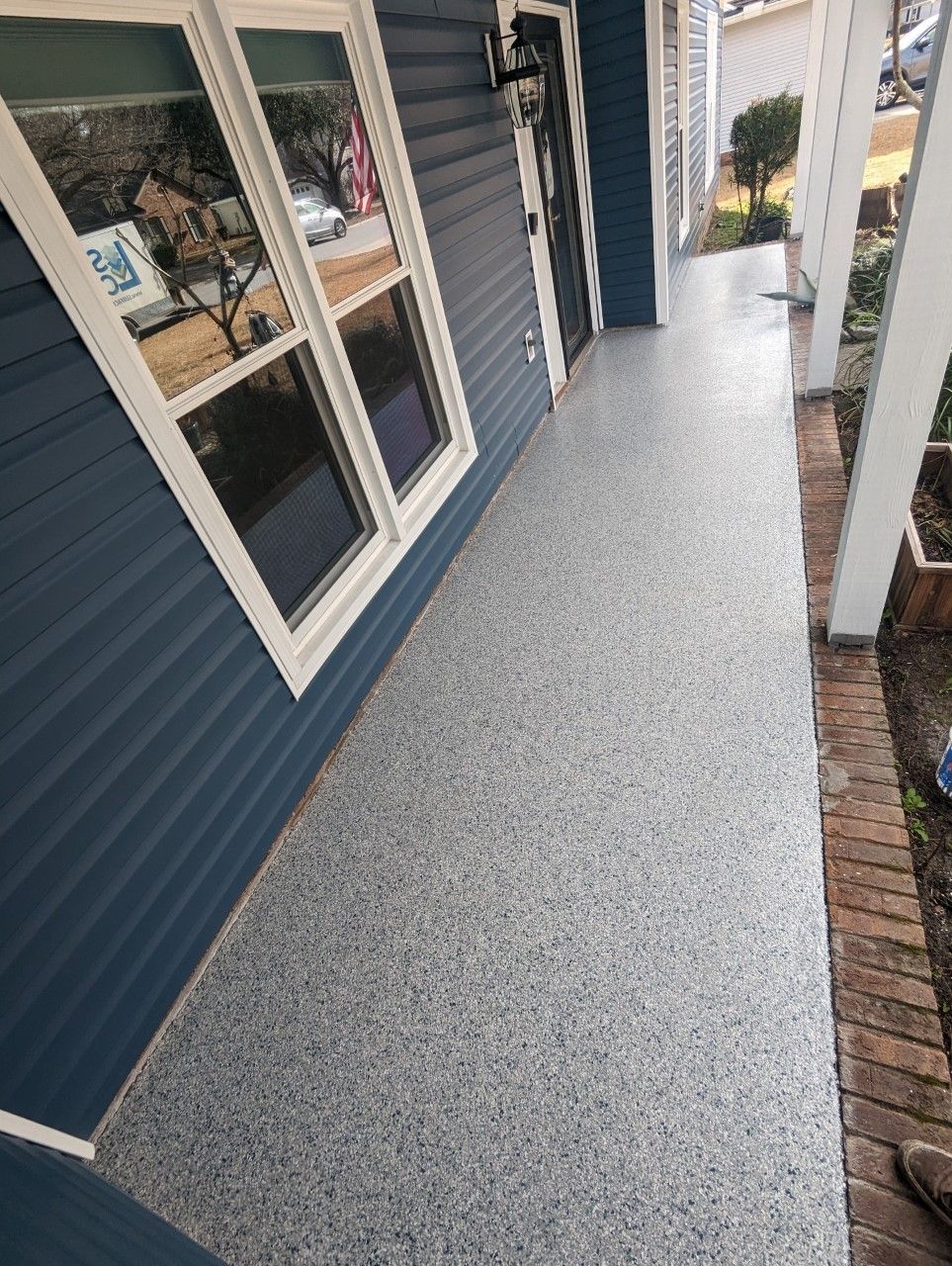 A narrow, gray speckled porch floor along a blue-sided house with white window frames. 
