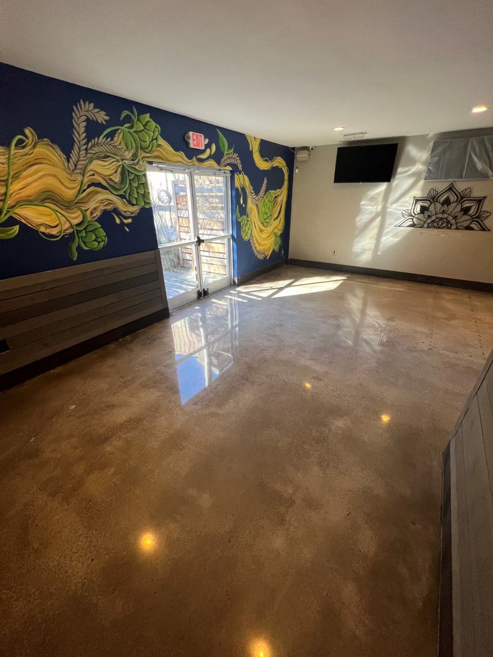 Empty room with a vibrant mural on the wall and glass doors leading outside. Polished concrete floor, sunlight.