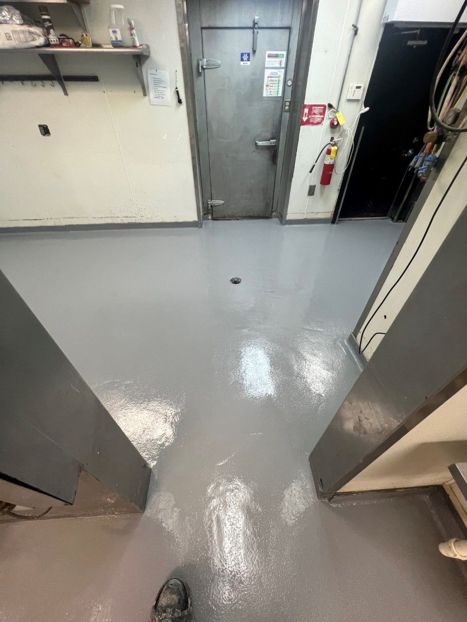 A gray floor in a commercial or industrial setting. A metal door, fire extinguisher, and two gray support columns.