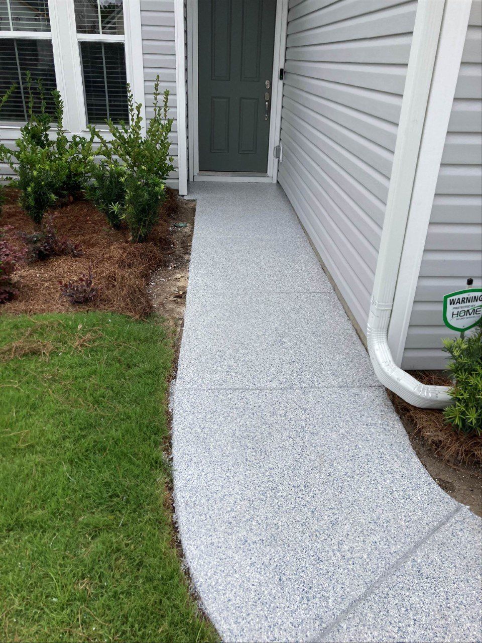 A gray speckled concrete walkway leads to a green door. The walkway is bordered by grass and a garden bed.