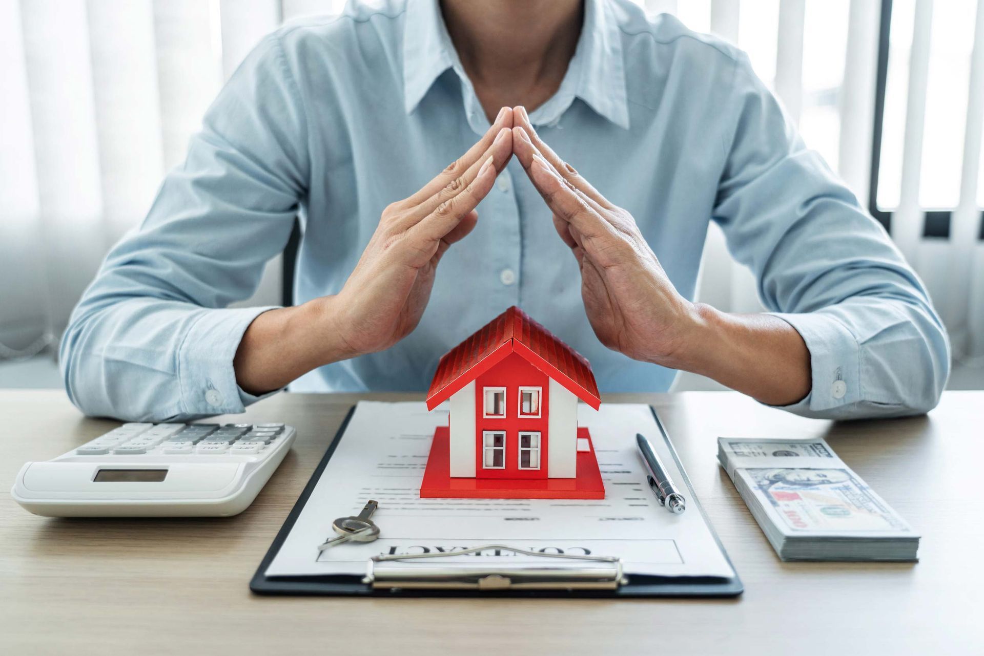 Hands forming a roof over a red house model on a clipboard, with calculator and cash on a desk