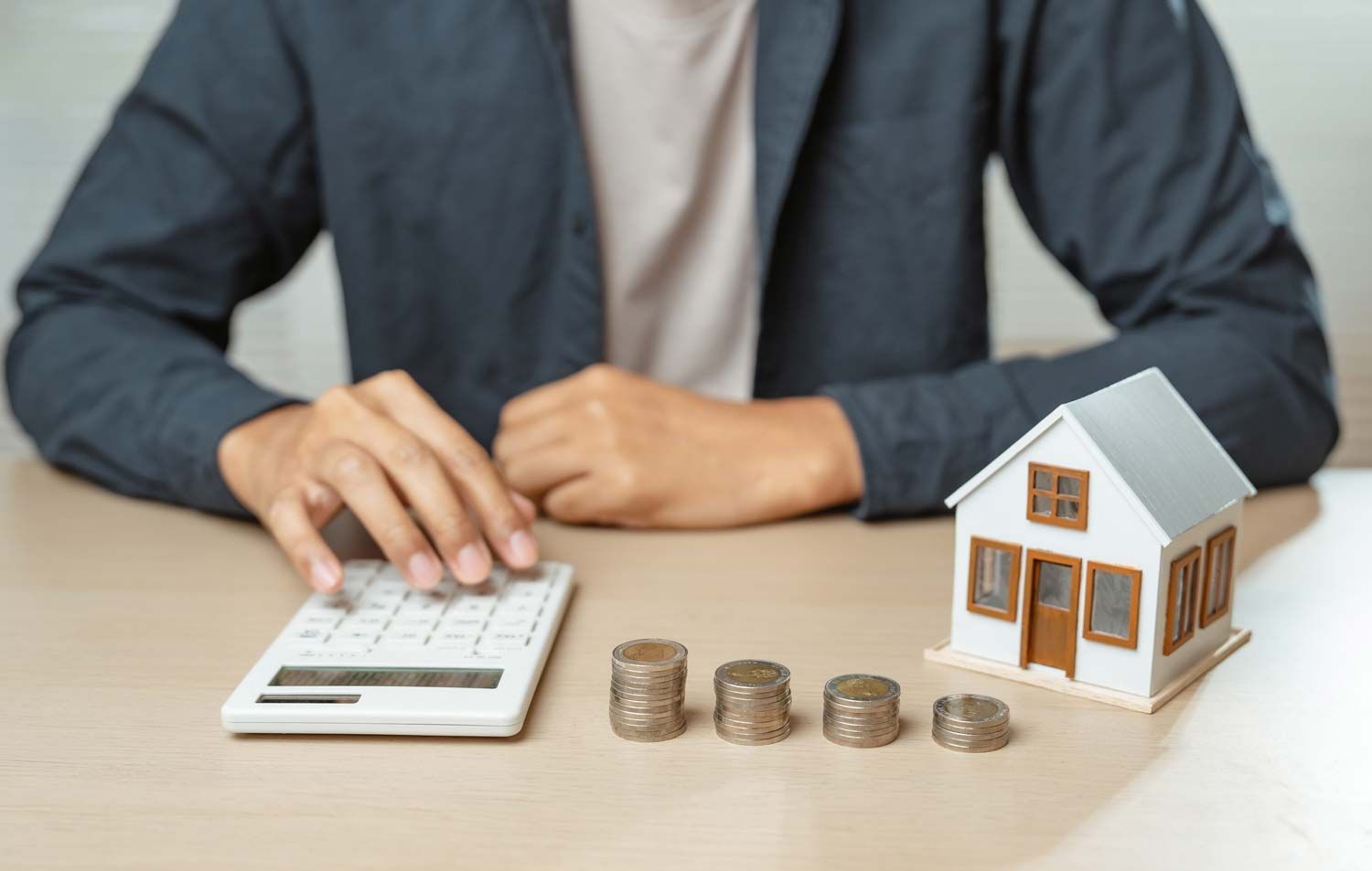 Person using a calculator beside house model and stacked coins on a desk, suggesting budgeting or mortgage planning