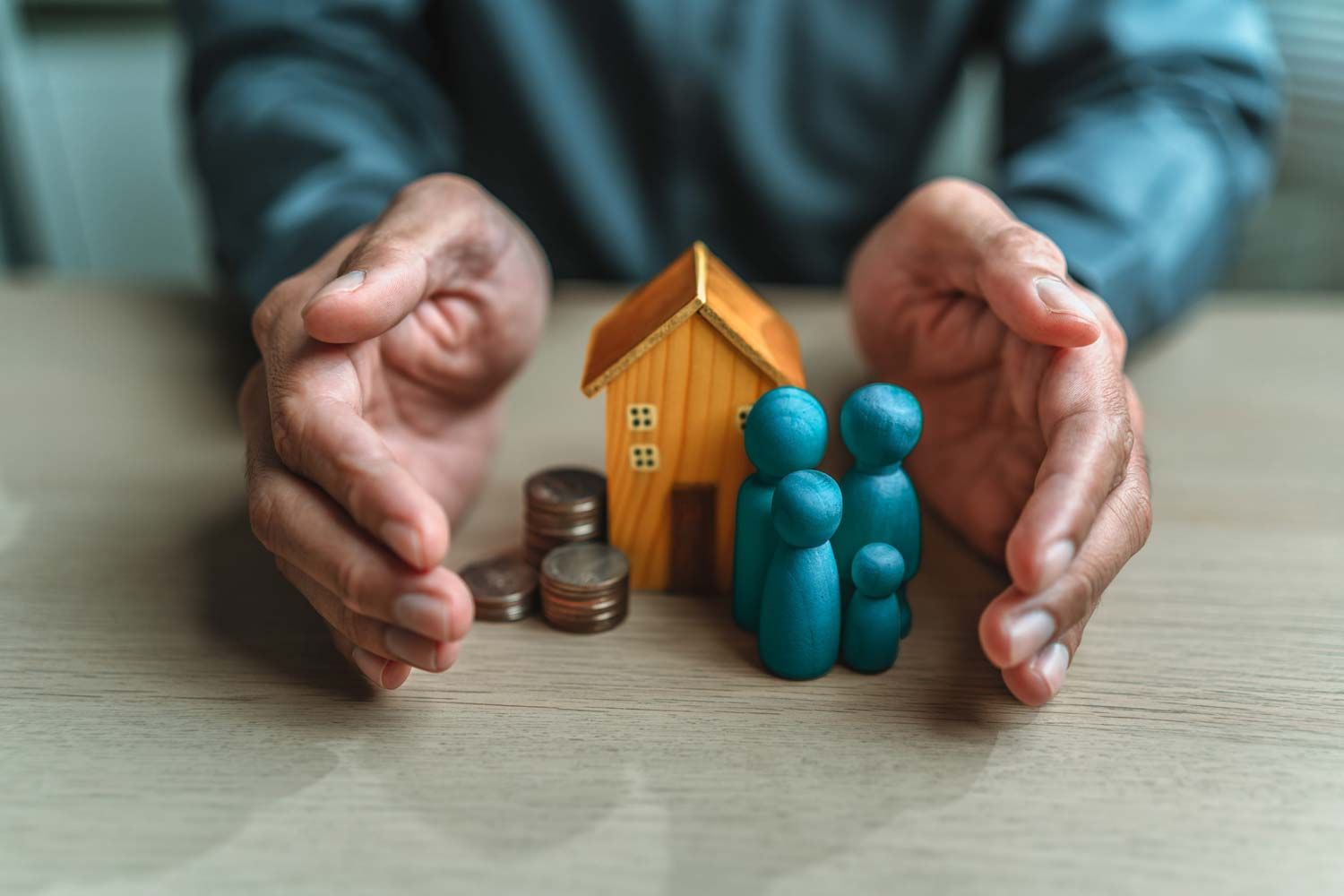 Hands cupping a small house, coins, and blue figurines on a table