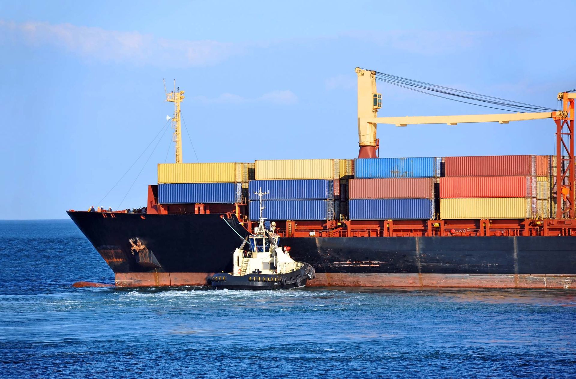 Cargo ship with stacked containers and a small tugboat on blue water under a clear sky