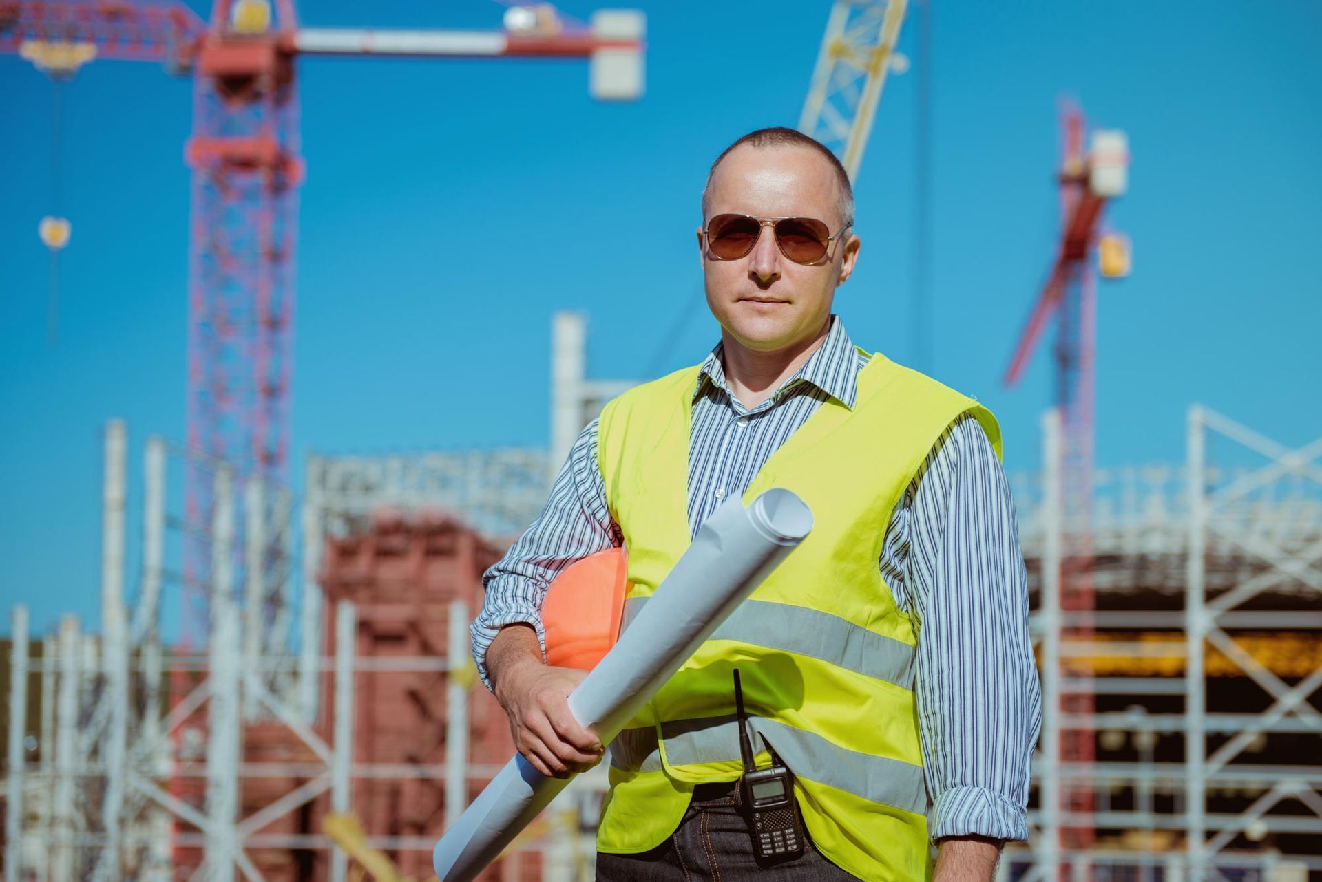 Construction worker in a neon vest holding rolled plans at a building site with cranes in the background