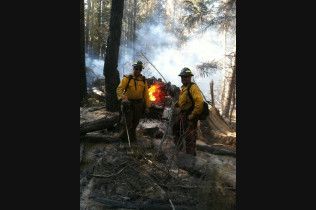 Firefighters in yellow gear stand near a smoky forest fire, with flames visible in the background