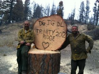 Two people stand beside a large tree stump carved “DJJG DC Trinity 2021” in a forest clearing.