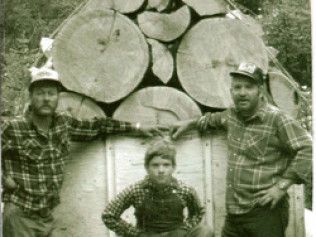 Three people pose in front of a large stack of cut logs outdoors.