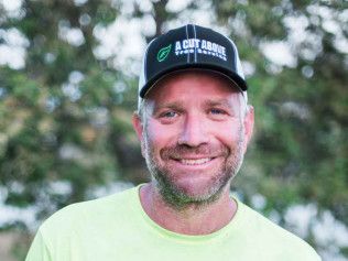 Smiling man in a neon yellow shirt and black cap outdoors with trees in the background