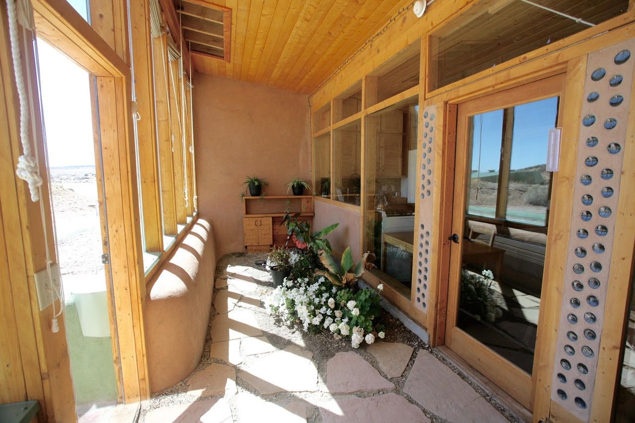 Interior of a tiny earthship greenhouse with South-facing windows, a bottle wall, and a few plants.