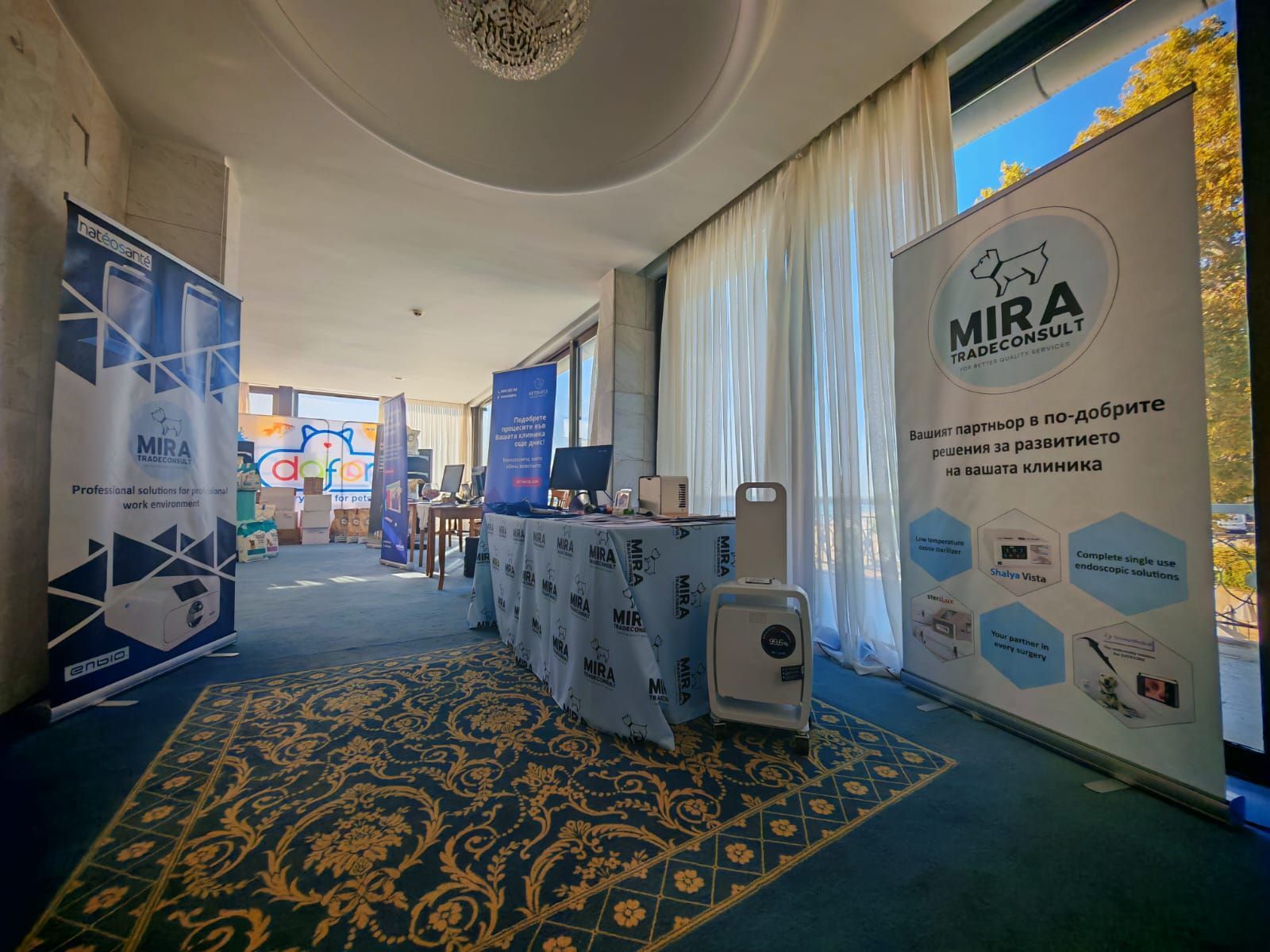 Щанд на търговско изложение с банери, рекламиращи „MIRA TRADECONSULT“. Маса с продукти и килим на пода.