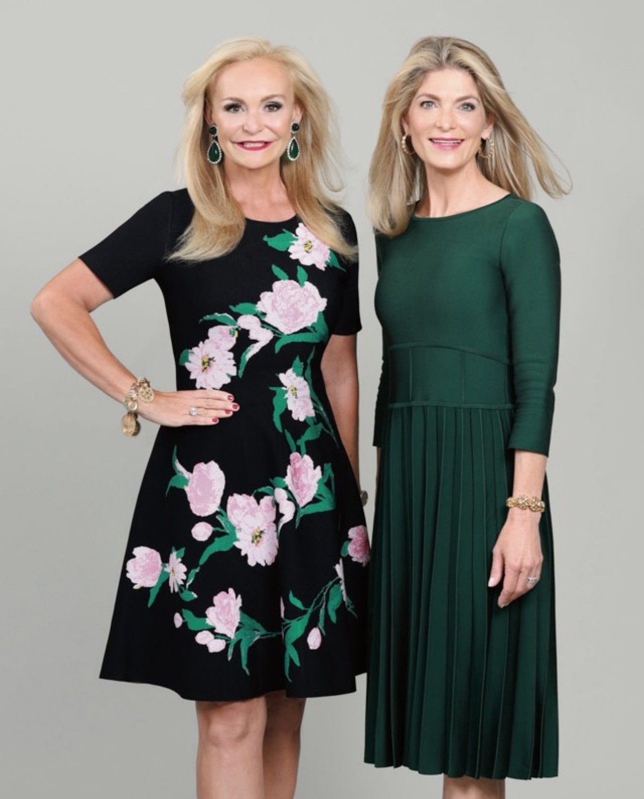 Two women stand side-by-side against a light gray background, one wearing a floral dress and one in a solid green dress.