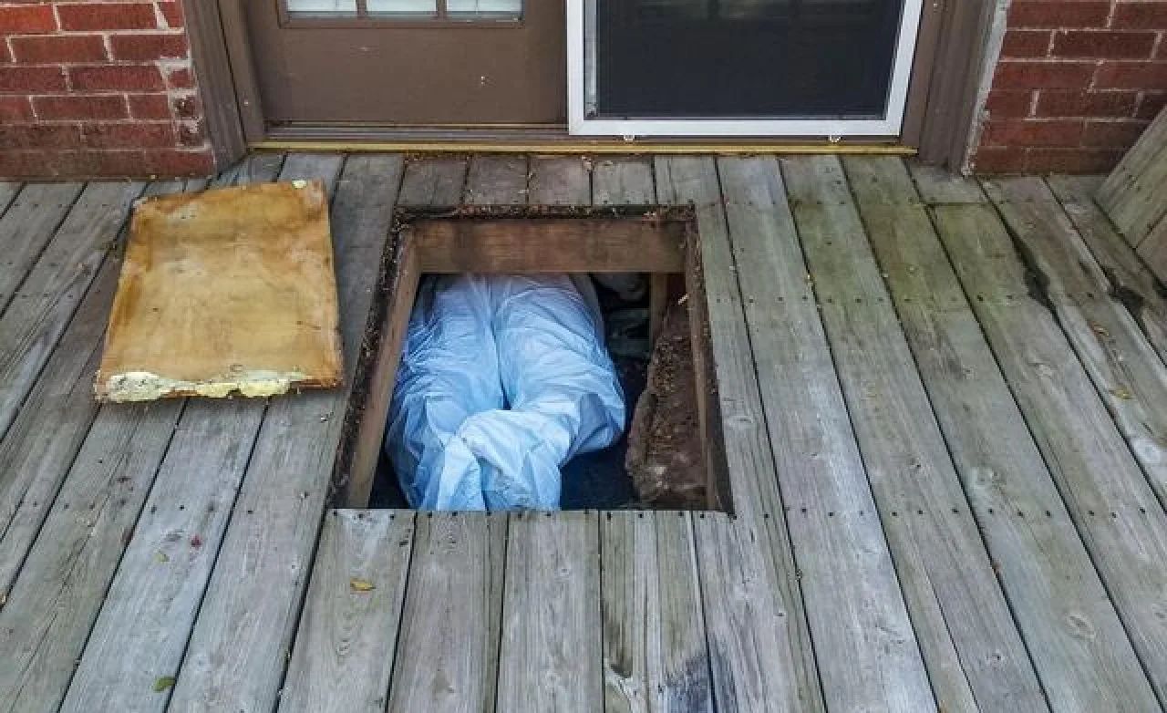 Person in blue clothing crawling through a cutout in a wooden porch floor near a door