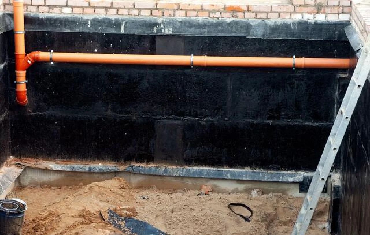 Black waterproofed wall with orange pipes along a basement trench and a ladder on the right