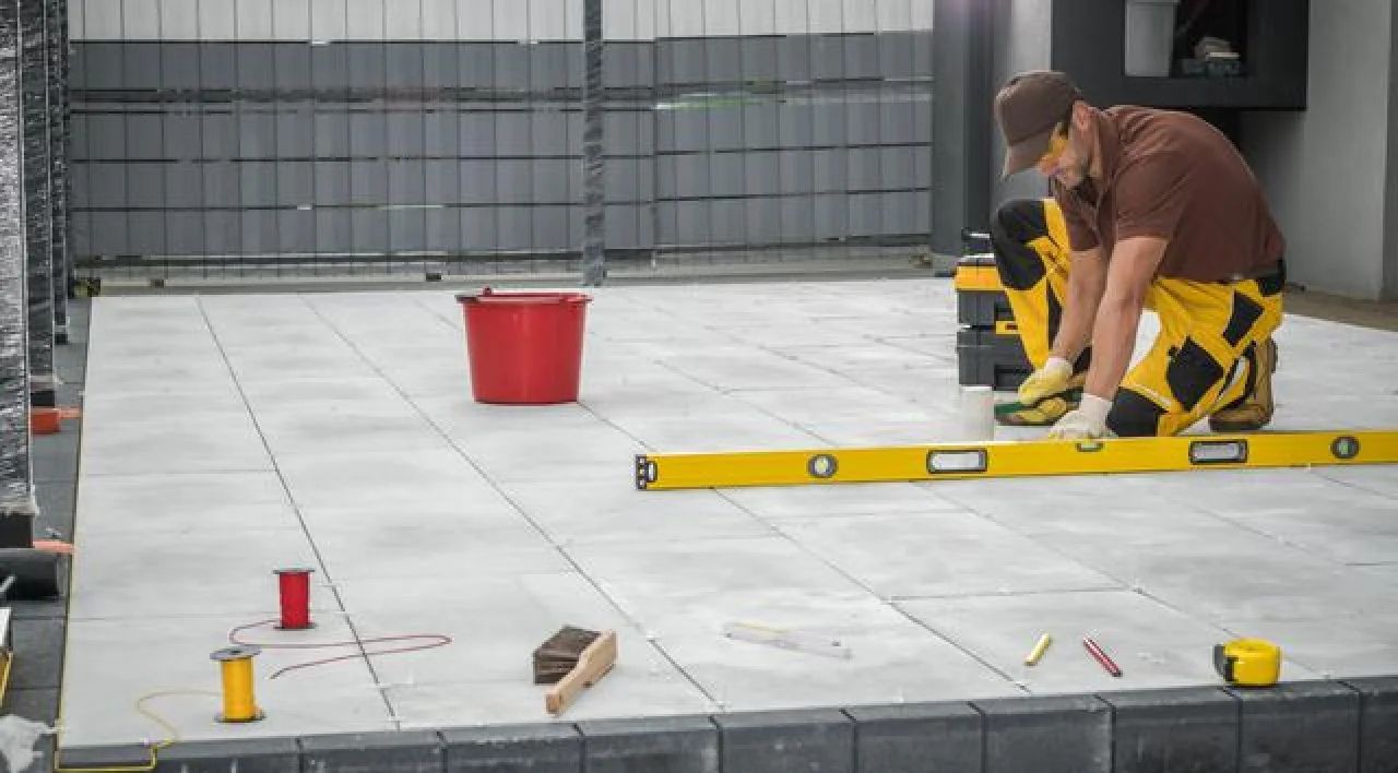 Worker installing tile on outdoor patio with red bucket and yellow level