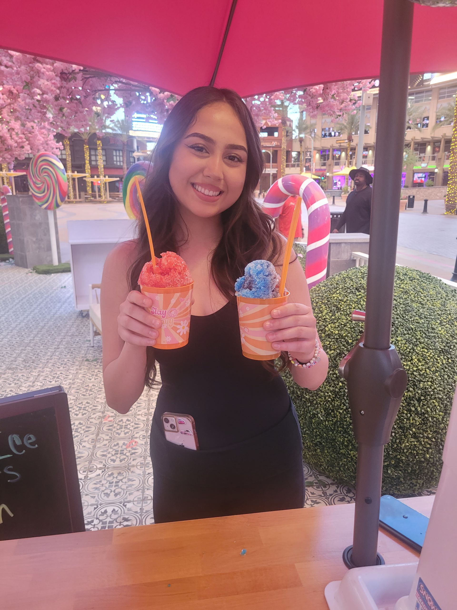 Woman holding two colorful slushies under a red umbrella. Outdoor setting with decorations.