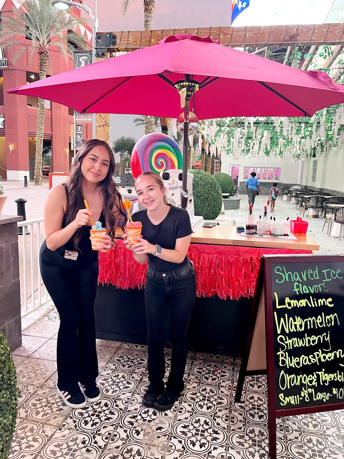 Two women smiling, holding drinks, beside a shaved ice stand. Pink umbrella, flavors sign.