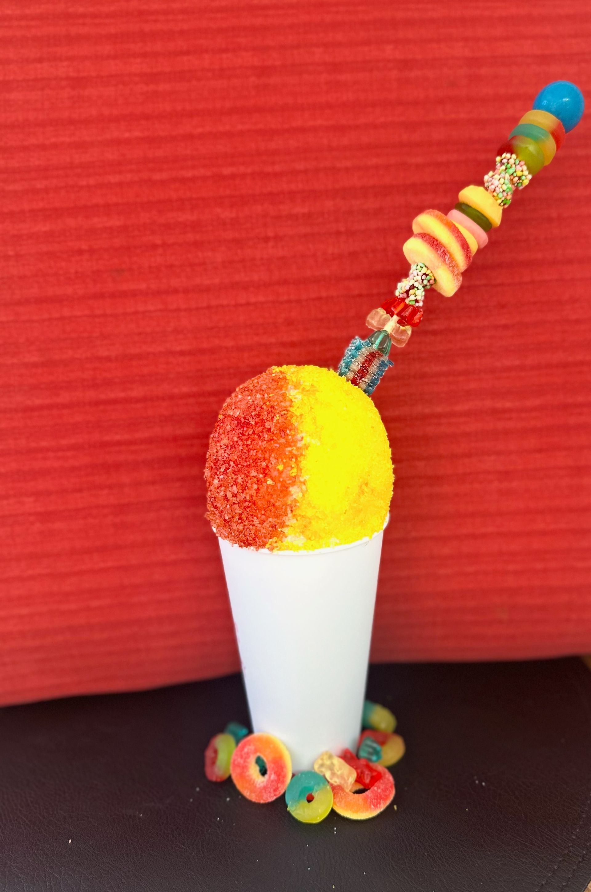 A colorful snow cone in a white cup, decorated with candy and a candy skewer, set against a red background.