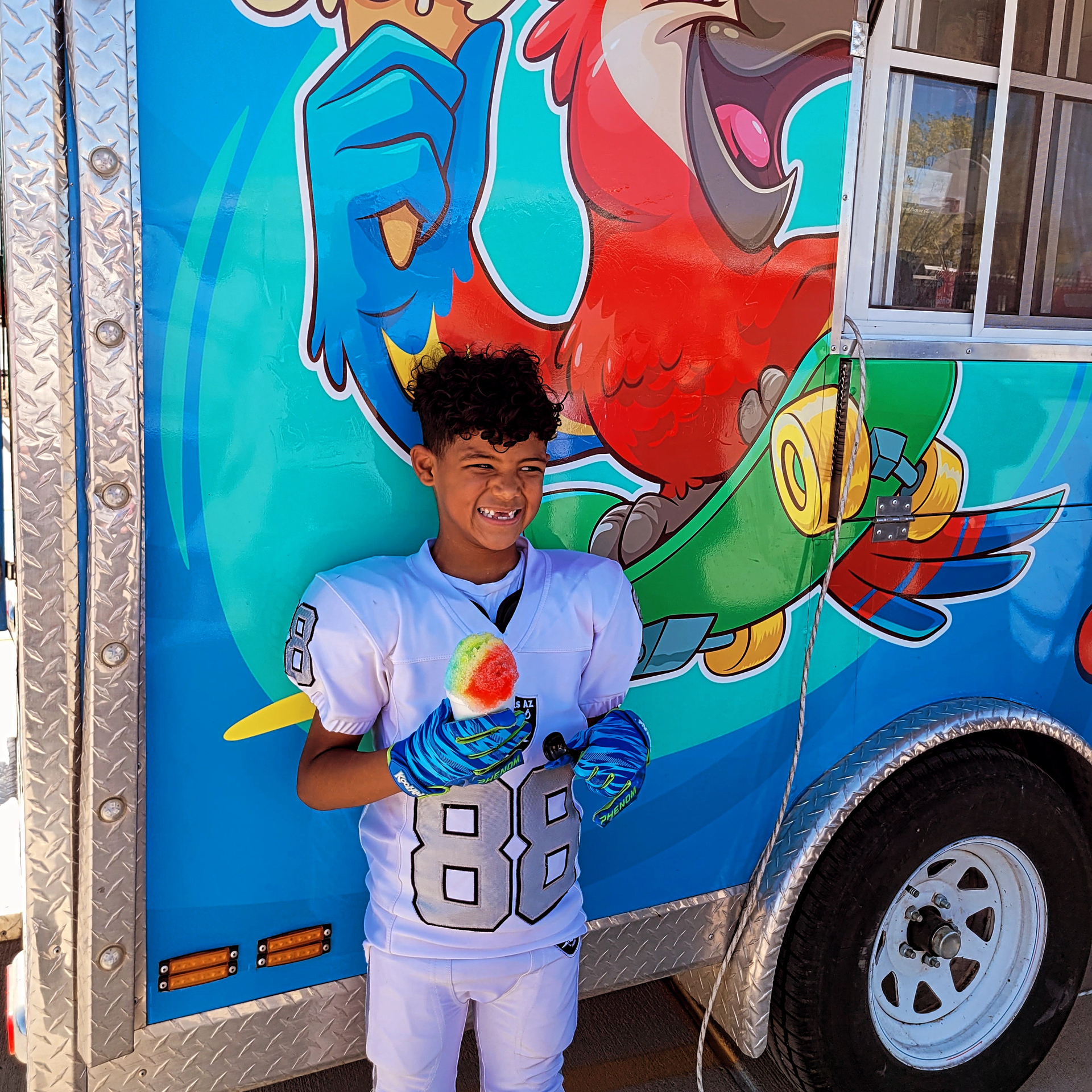 Young person in football uniform holding a treat by a colorful food truck with a parrot mural.