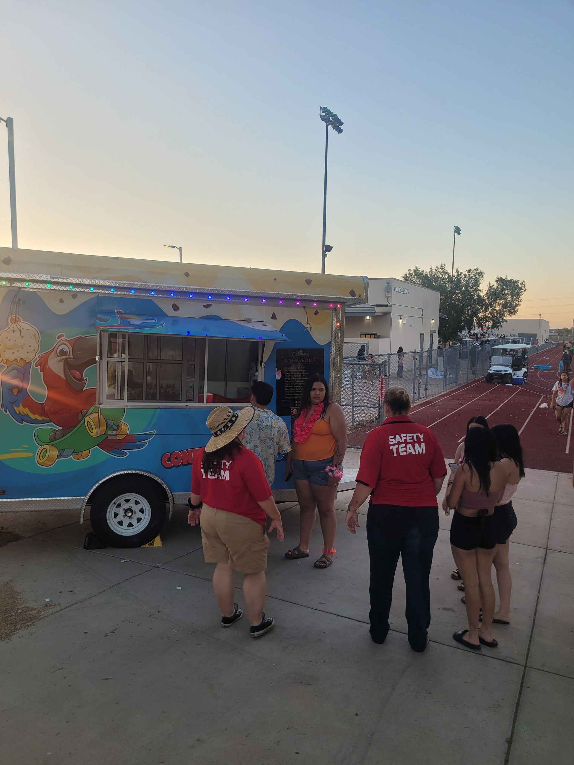 Food truck with people, some wearing red shirts, near a track at dusk.