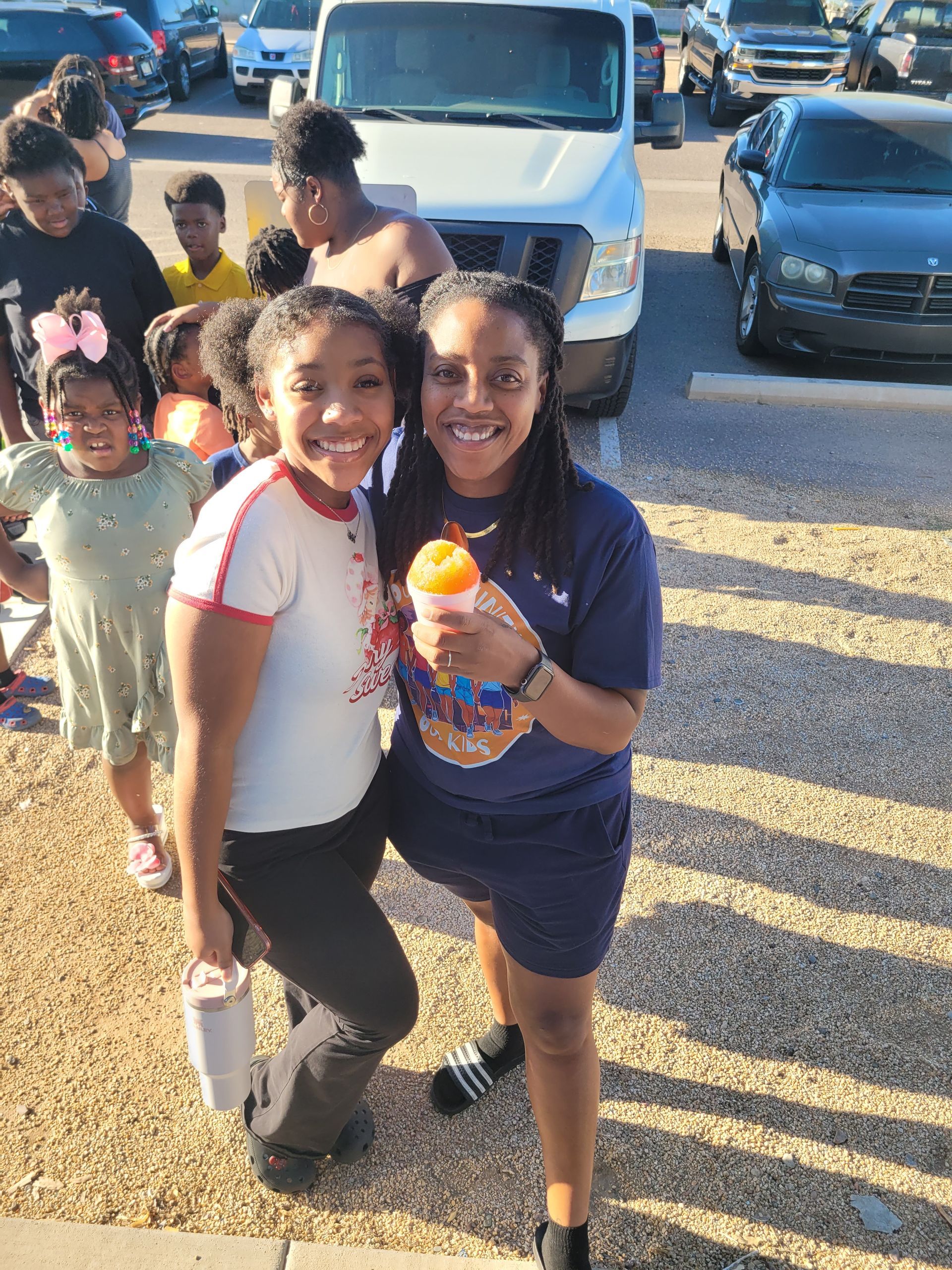 Two smiling people holding snacks pose outdoors with a group of people and vehicles.
