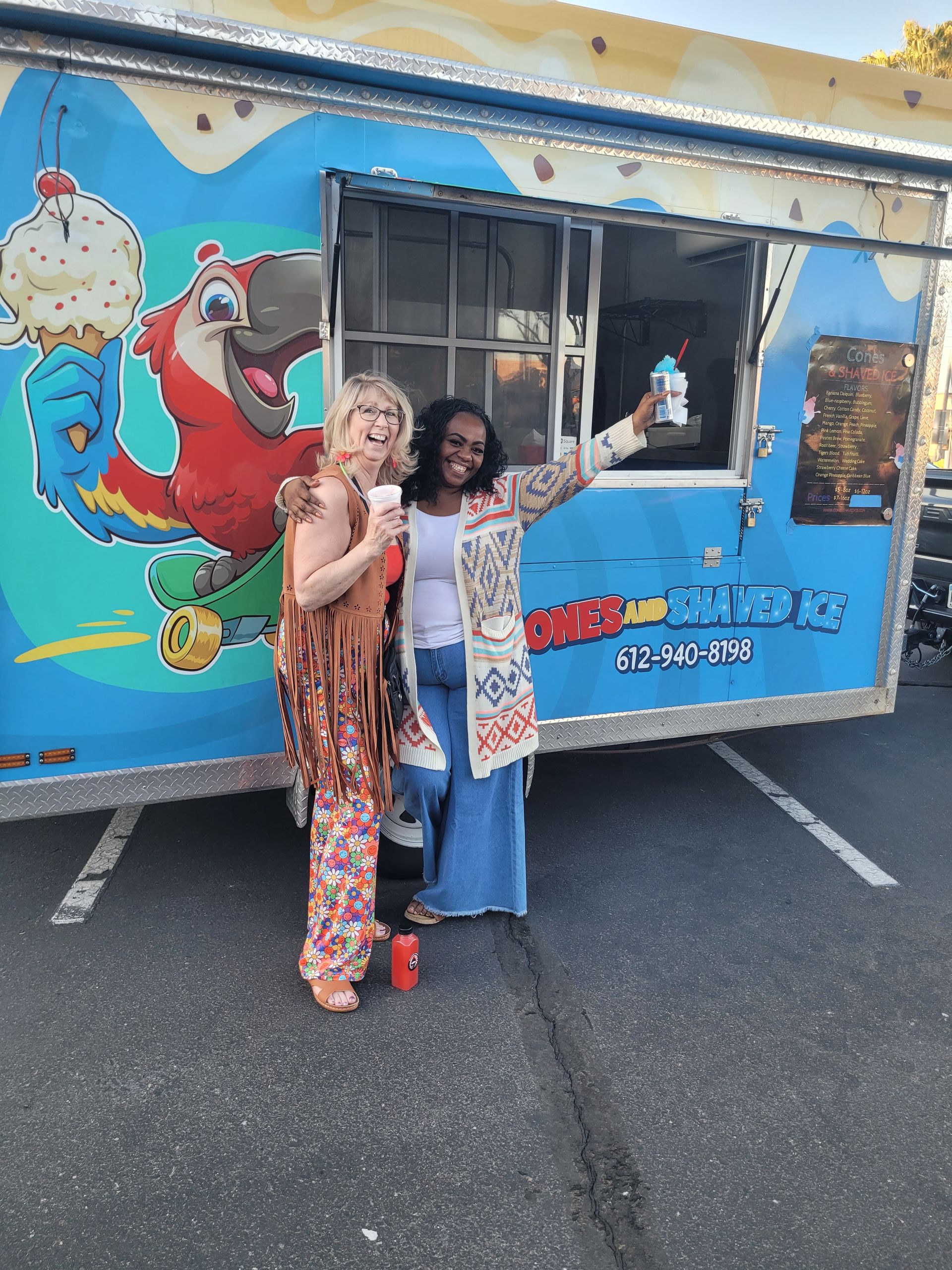 Two women smiling, posing with ice cream in front of a colorful ice cream truck with a parrot graphic.