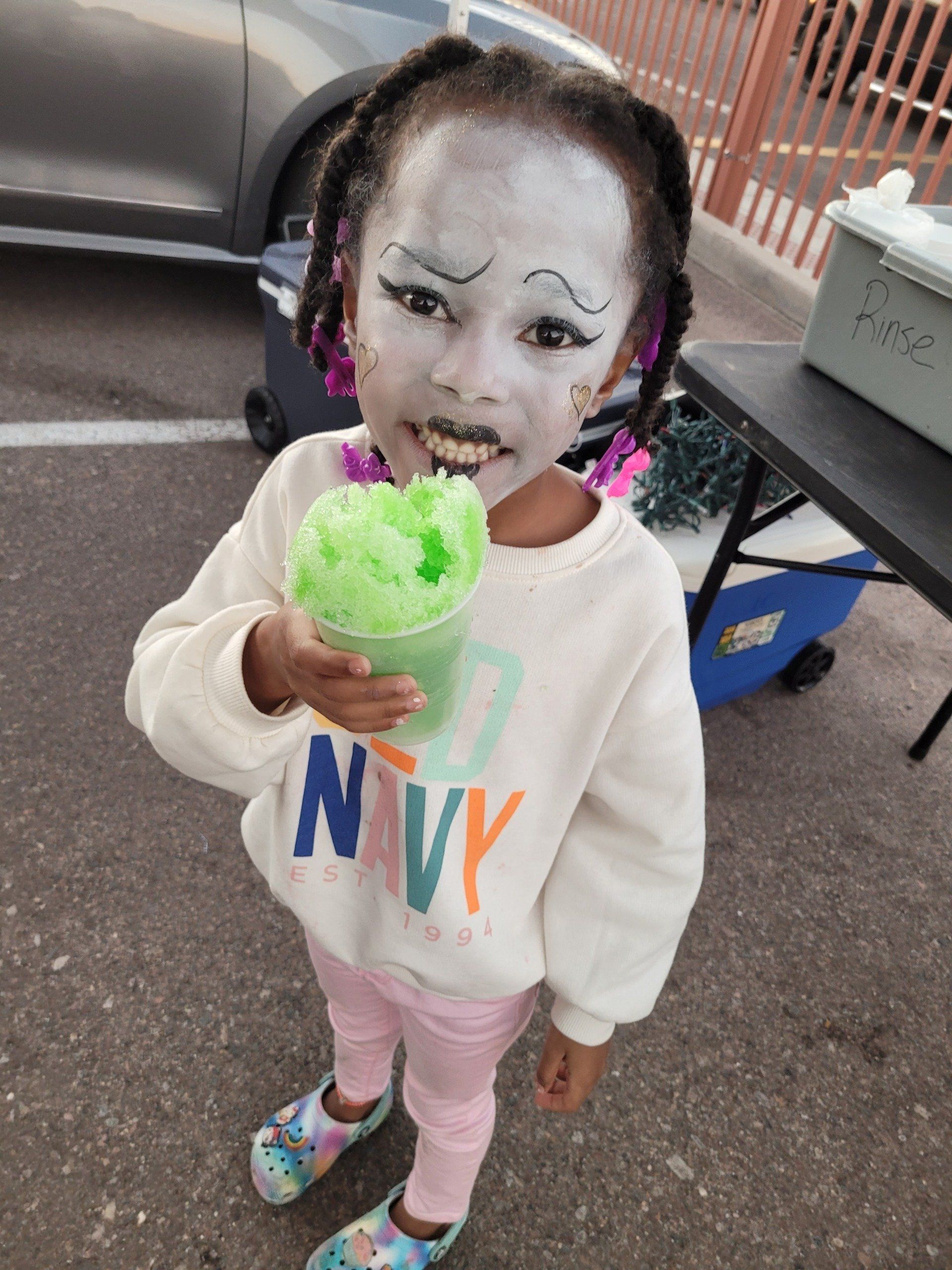 Child with face paint, holding a green snow cone. Wearing a cream sweatshirt, pink leggings, and colorful shoes. Outdoors.