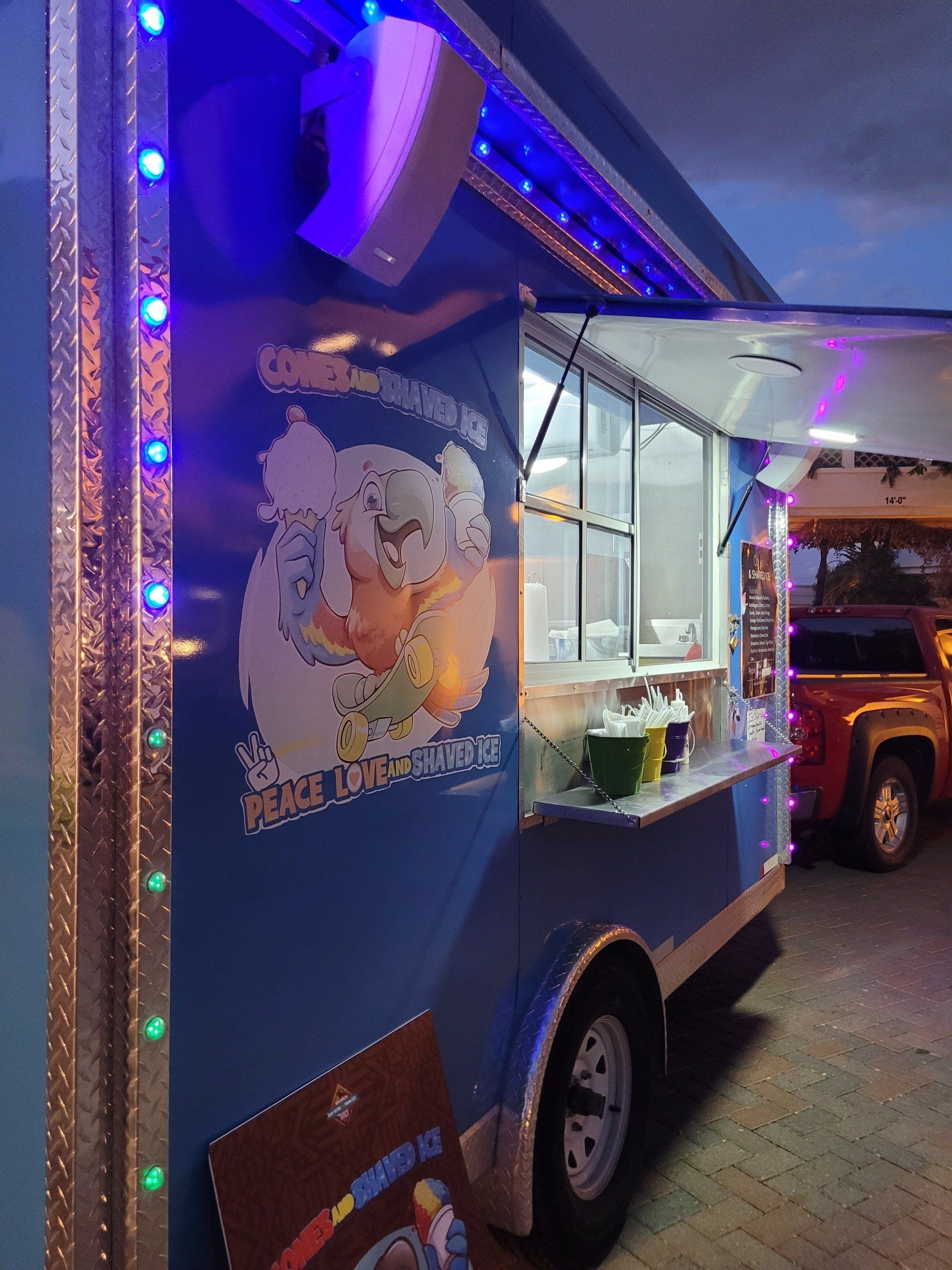 Blue food truck with open window, illuminated by blue lights, logo on side, parked next to a red truck.