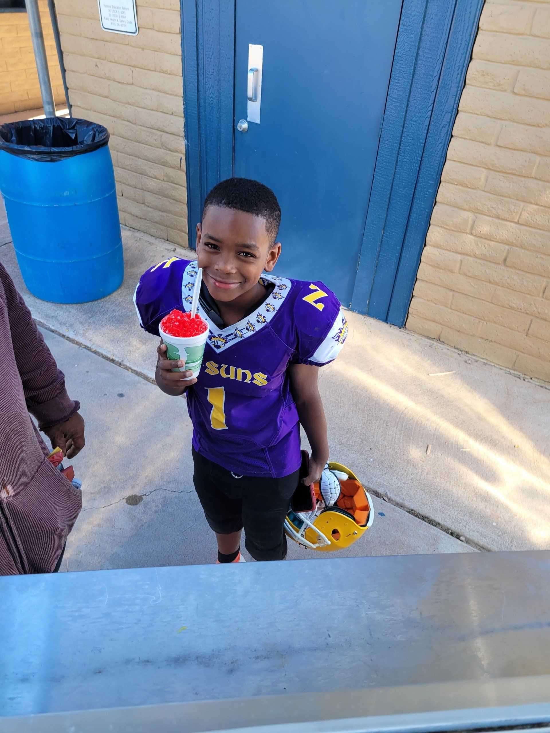 Young football player holding a snow cone. Purple and gold jersey, helmet, outside by a blue door.