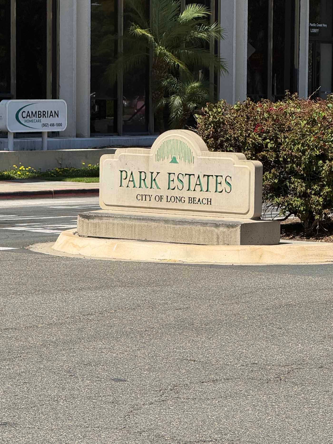 Park Estates entrance sign beside a driveway, with landscaped shrubs and a building in the background.