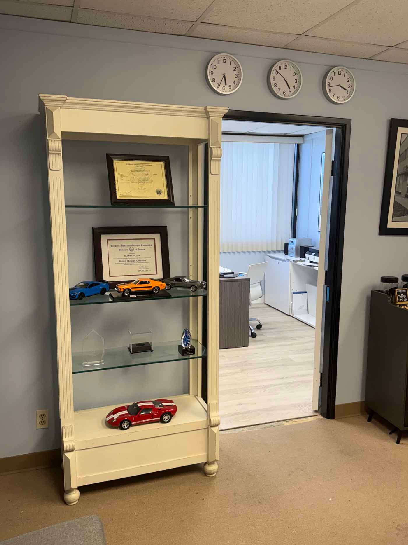 Tall white display cabinet with awards and toy cars in an office hallway beside an open doorway to a workroom