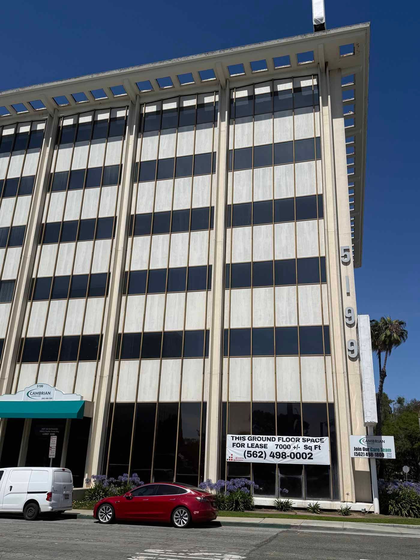Tall white office building with vertical windows, a red car, and a white van parked in front.