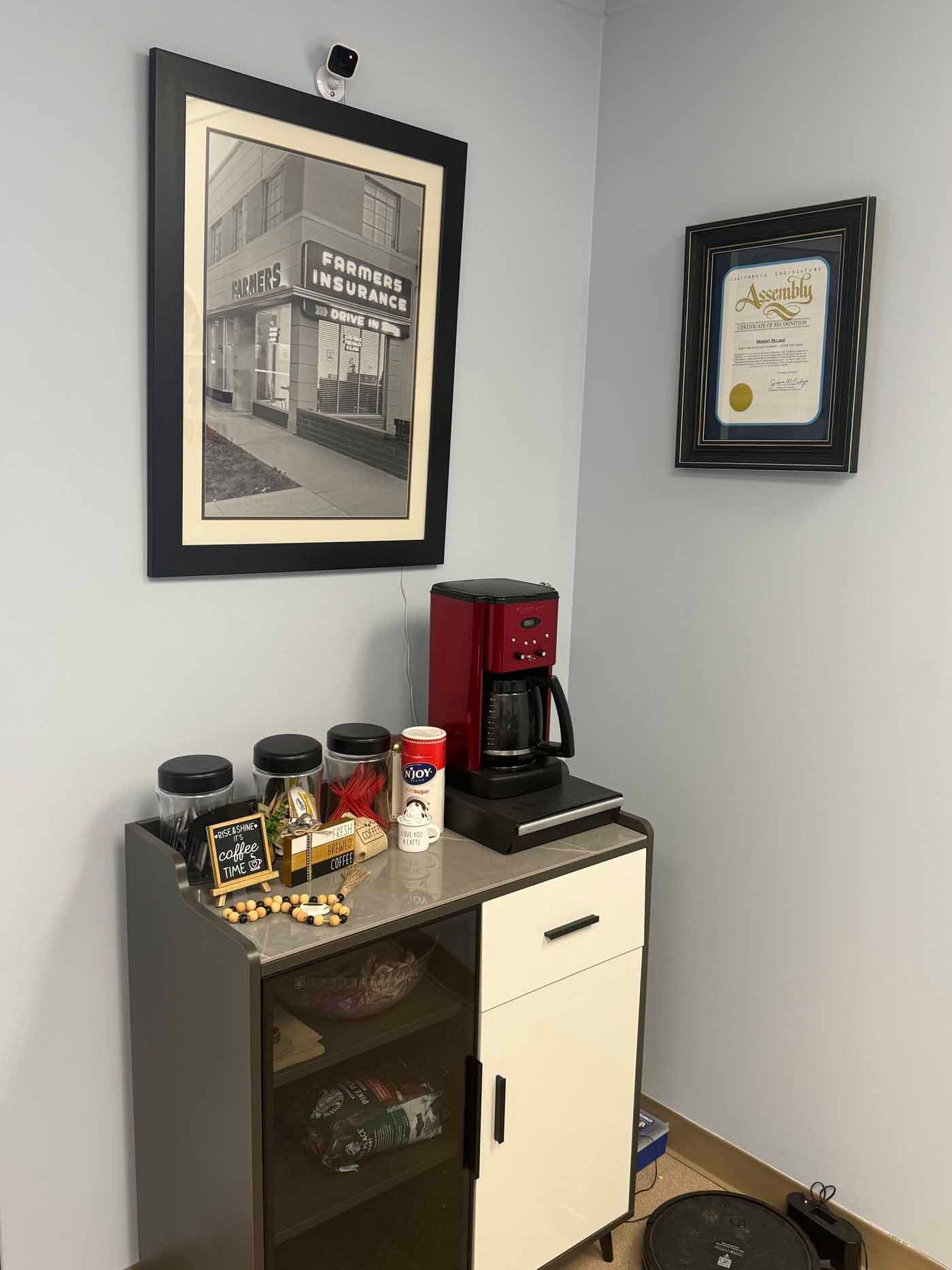 Coffee station with red espresso machine, mugs, snacks, and framed wall art in a corner cabinet.