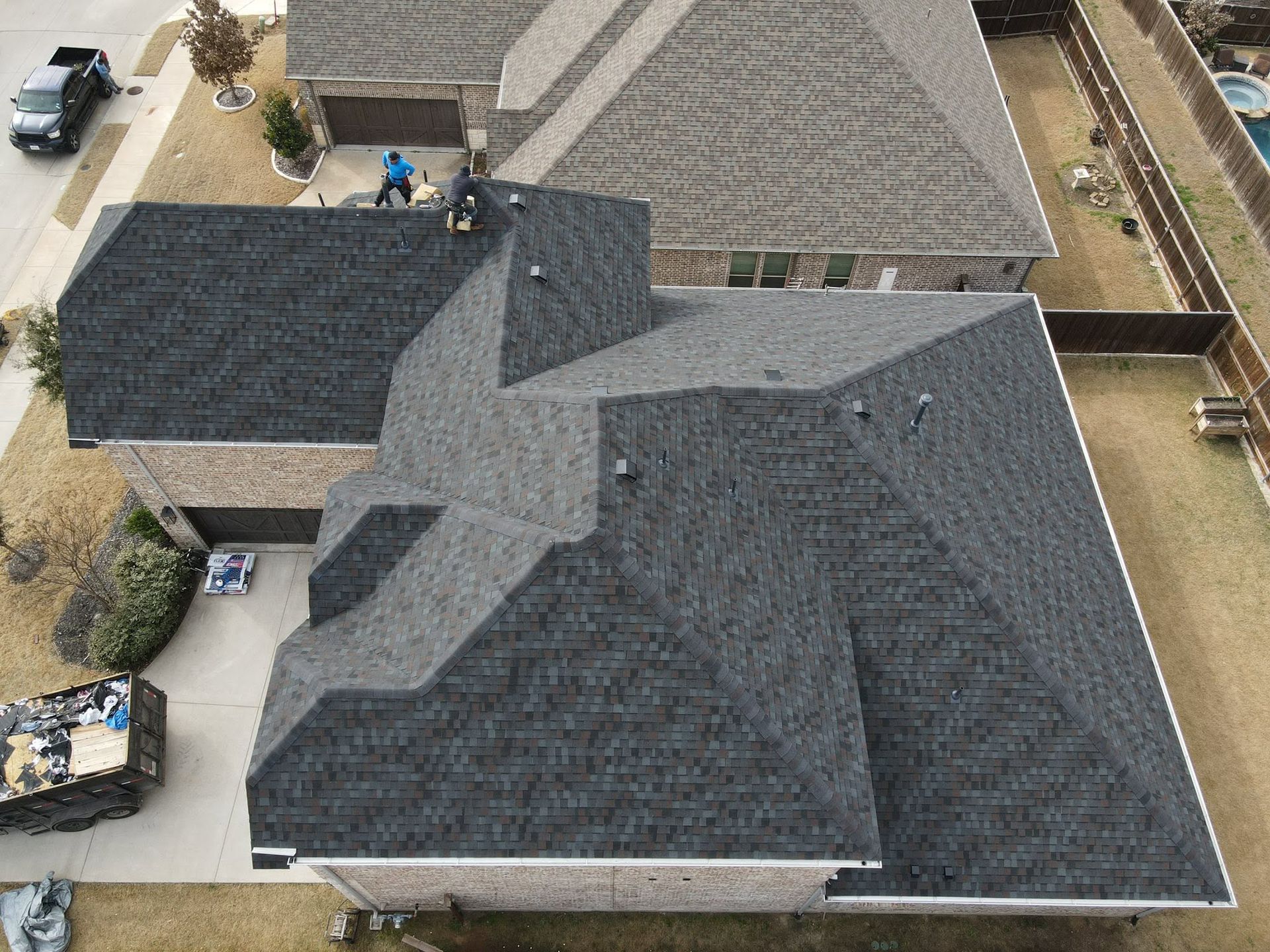 Aerial view of a suburban house with dark gray roofs and a driveway beside neighboring homes.